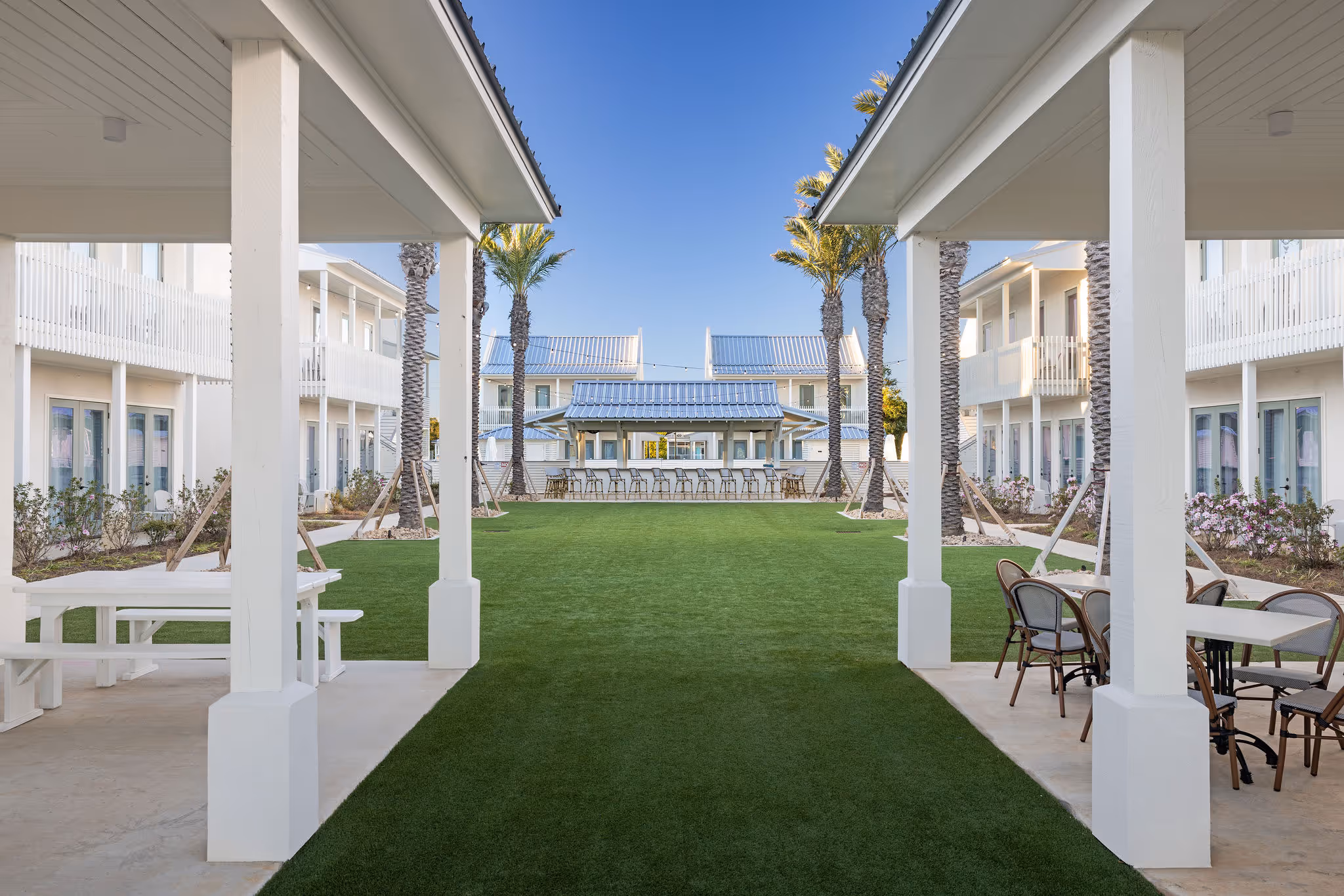 Outdoor courtyard with green grass, white buildings, palm trees, and covered seating areas with tables and chairs.