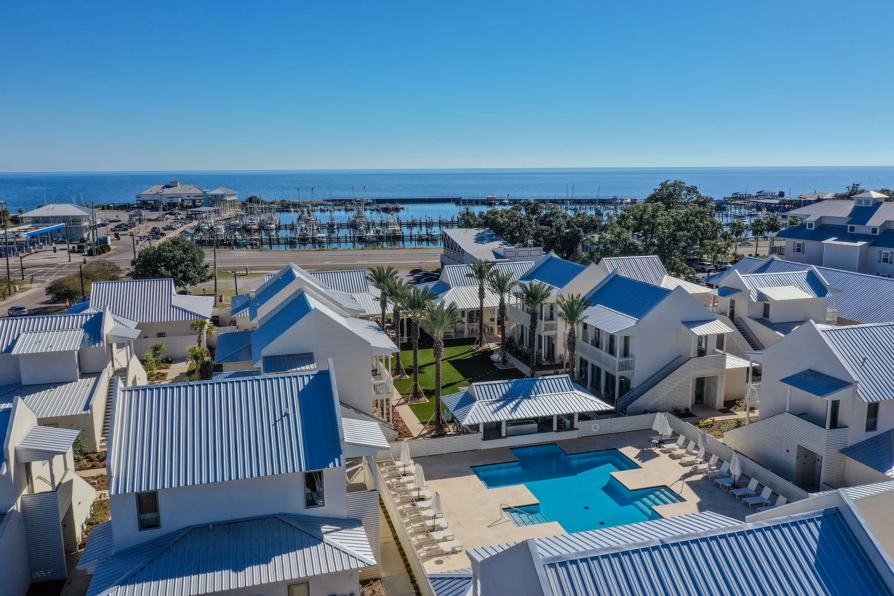 Aerial view of a coastal resort with white buildings, a swimming pool, palm trees, a marina, and the ocean in the background under a clear blue sky.