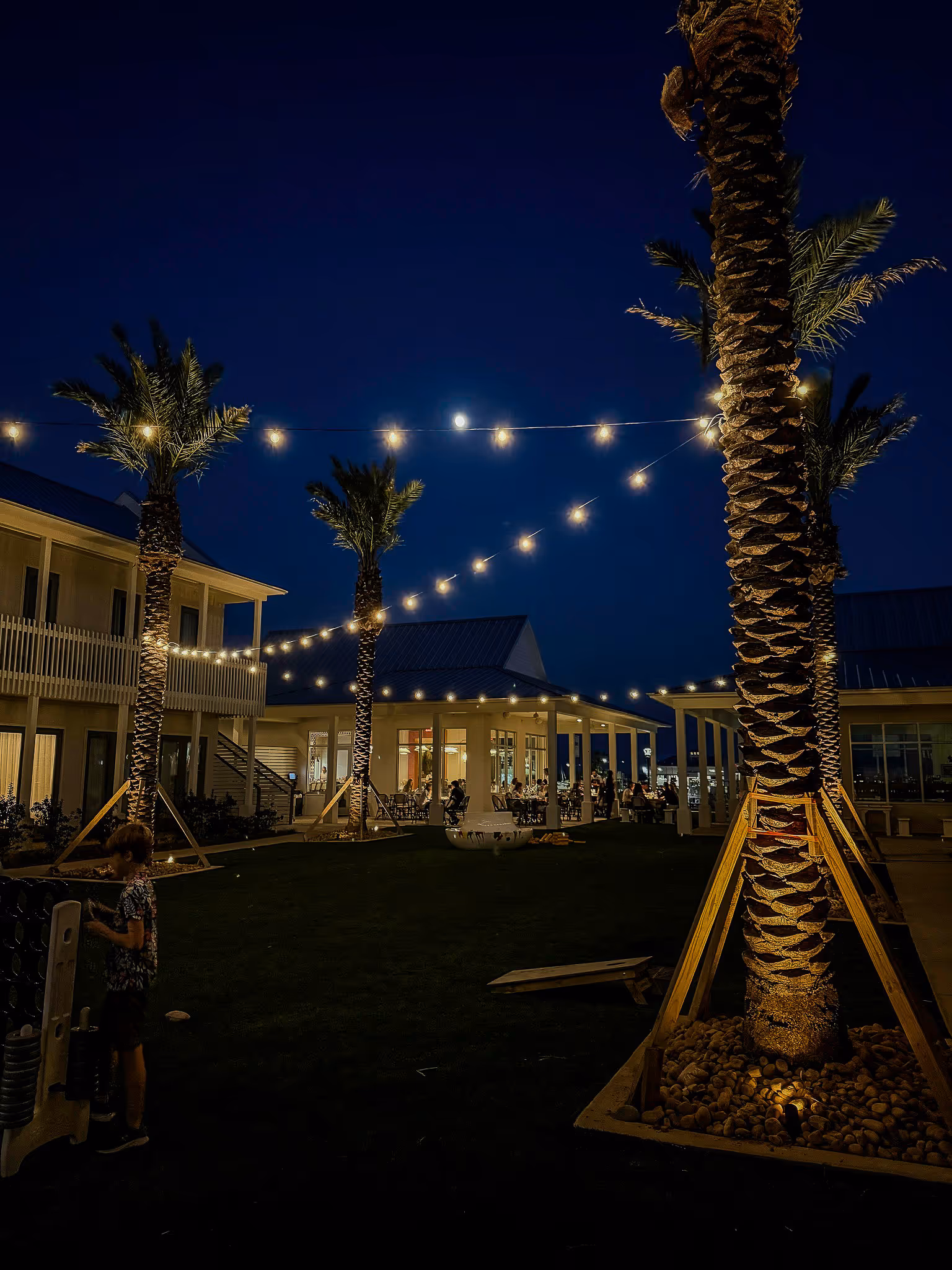 Outdoor night scene with string lights hanging between illuminated palm trees and people dining under a covered patio.
