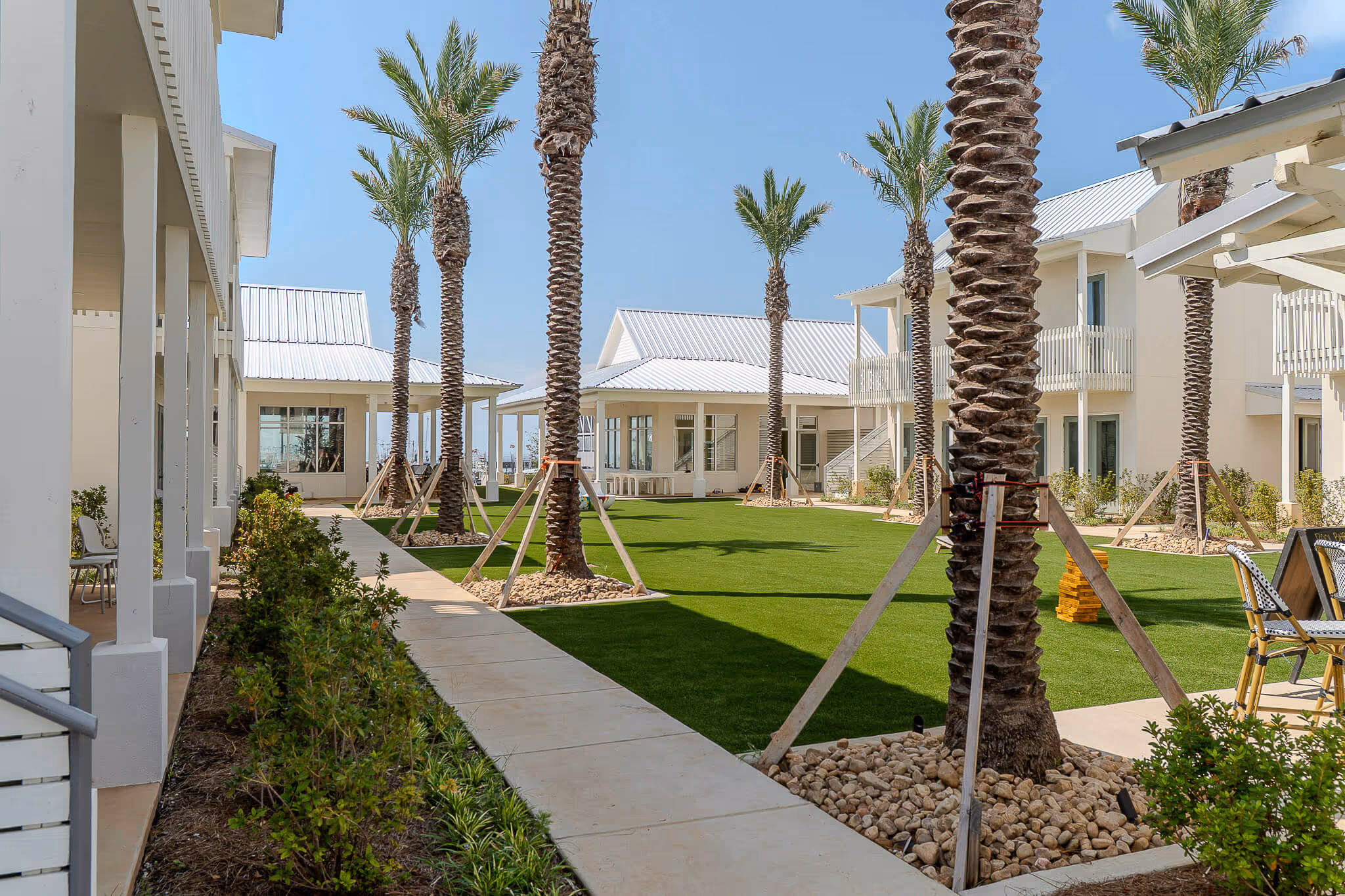 Modern residential courtyard with tall palm trees supported by braces, green lawn, and white buildings under a clear blue sky.