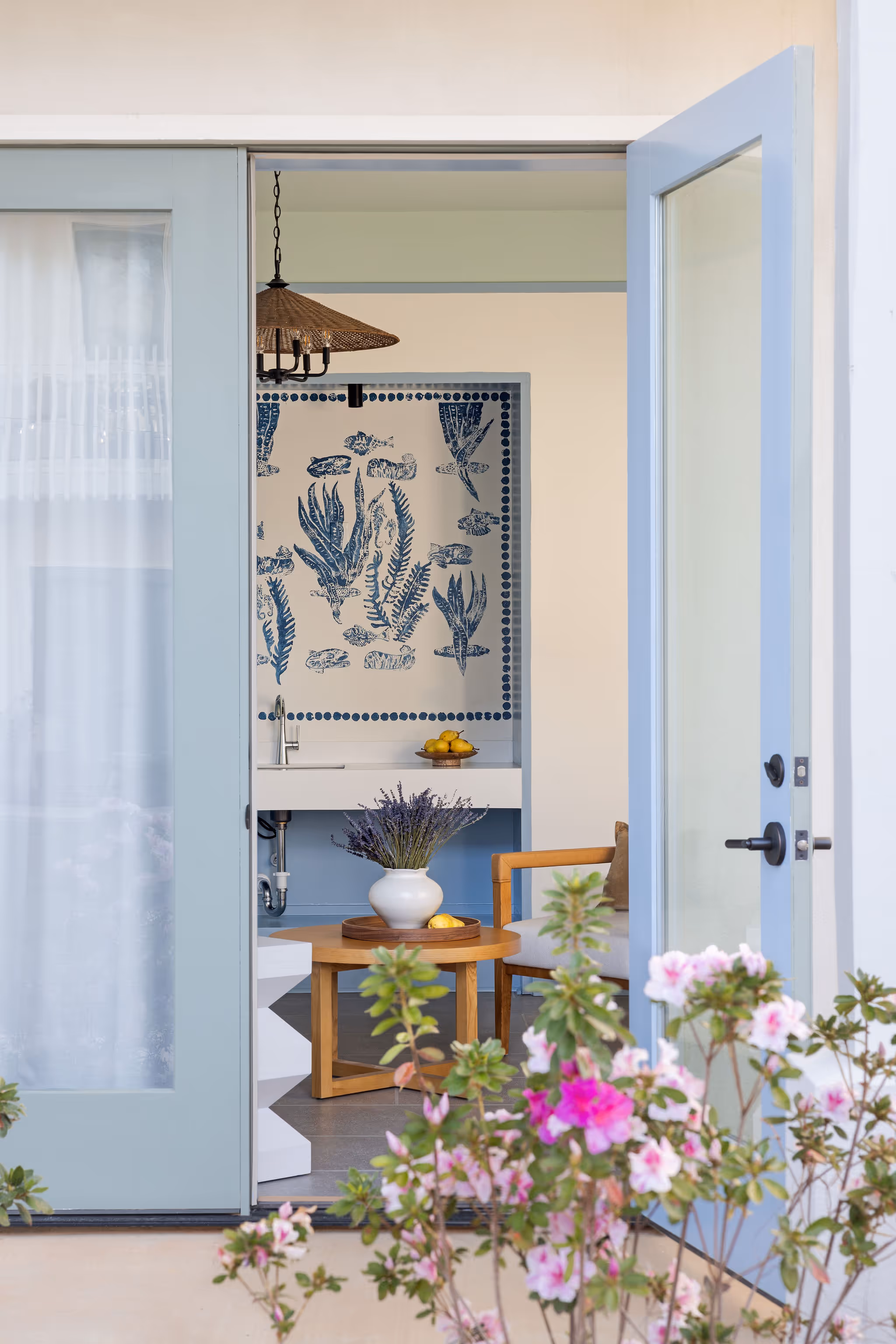 Open light blue door revealing a room with a wooden table holding a vase of lavender, a chair, and a wall with blue botanical art.