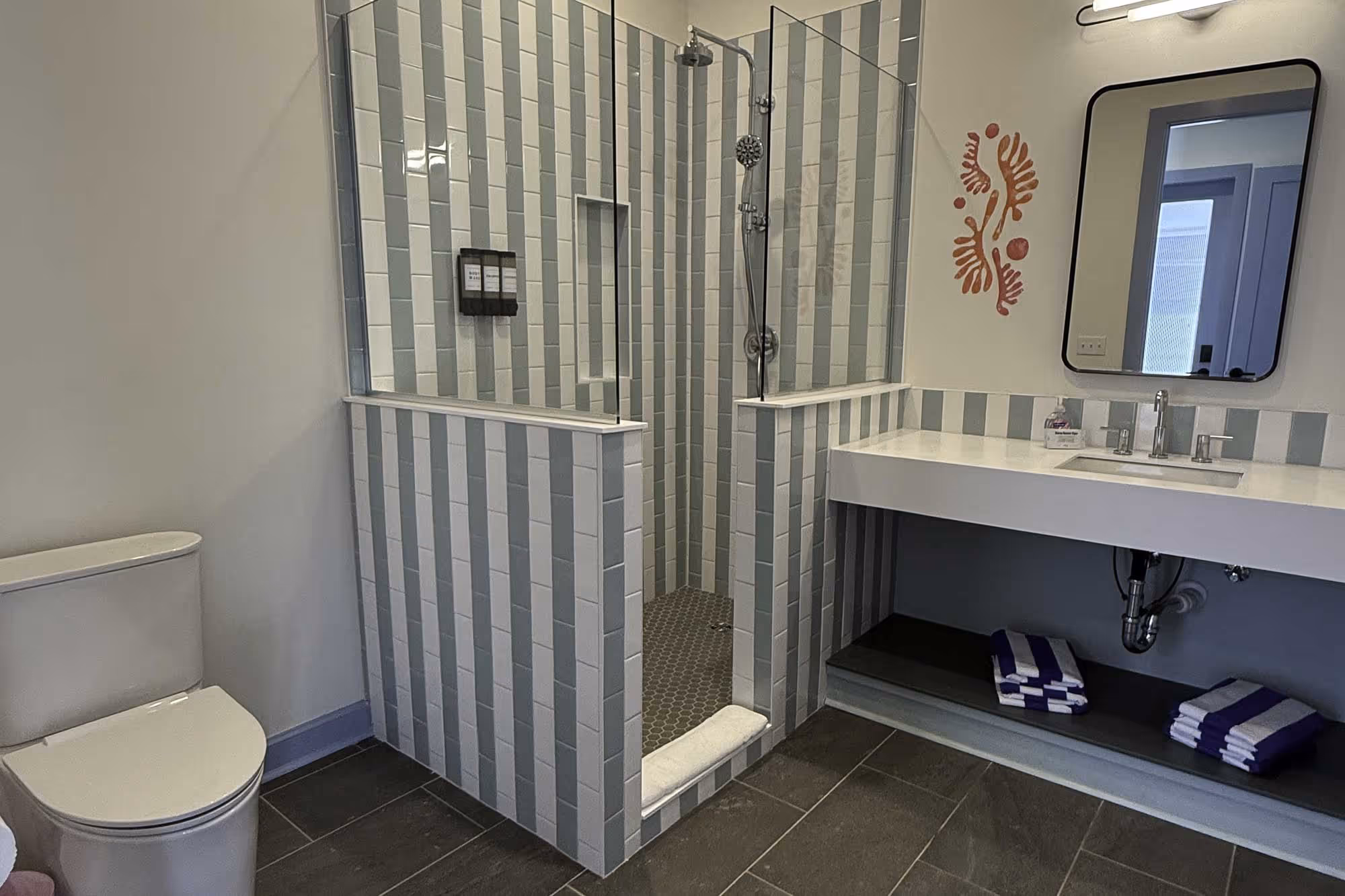 Modern bathroom with gray tile floor, open shower with blue and white vertical striped tiles, white toilet, and a white sink with mirror and folded blue and white towels underneath.