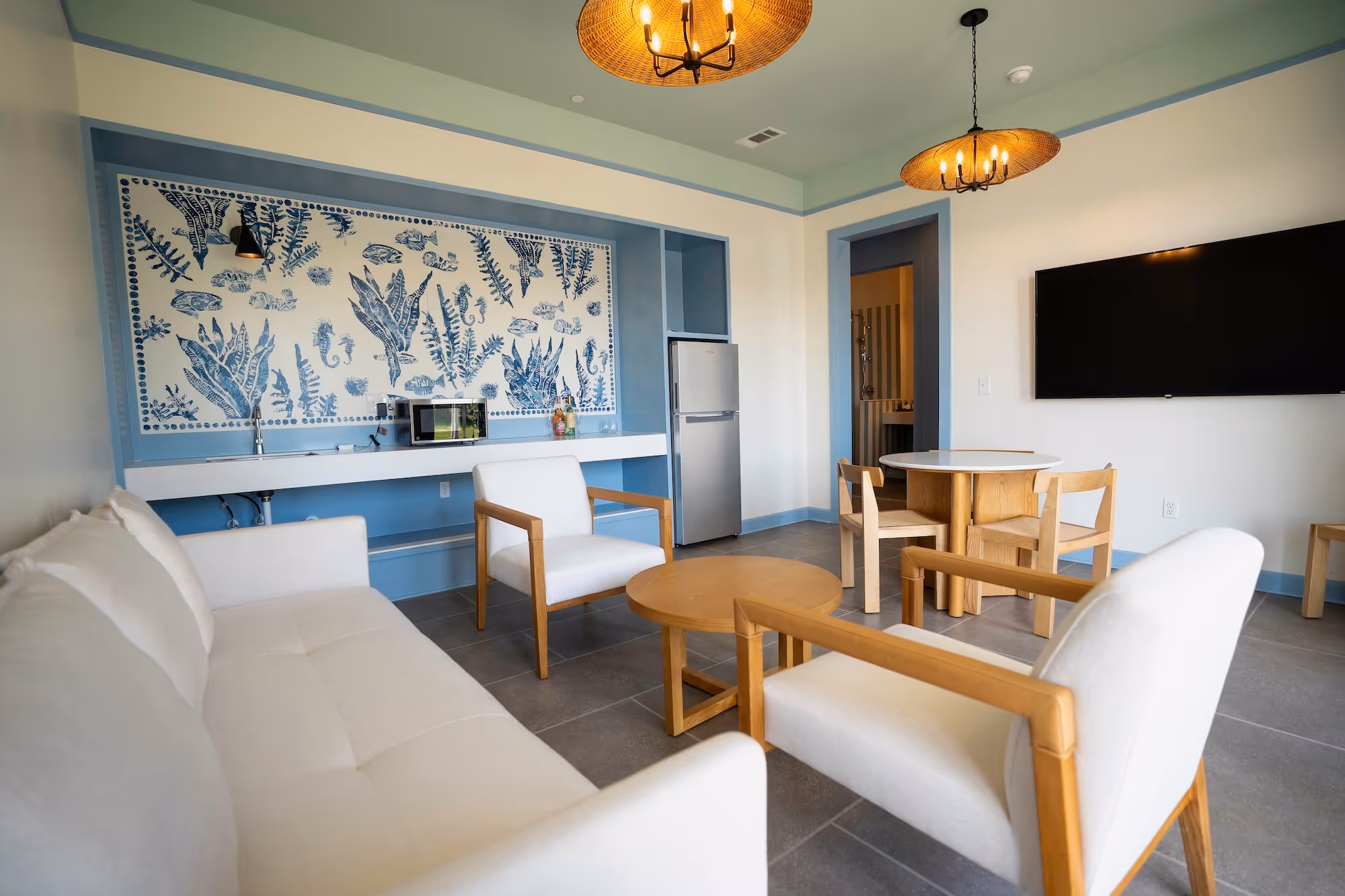 Modern living room with white cushioned wooden chairs, a sofa, round wooden coffee table, wall-mounted TV, and kitchenette with patterned blue and white backsplash.