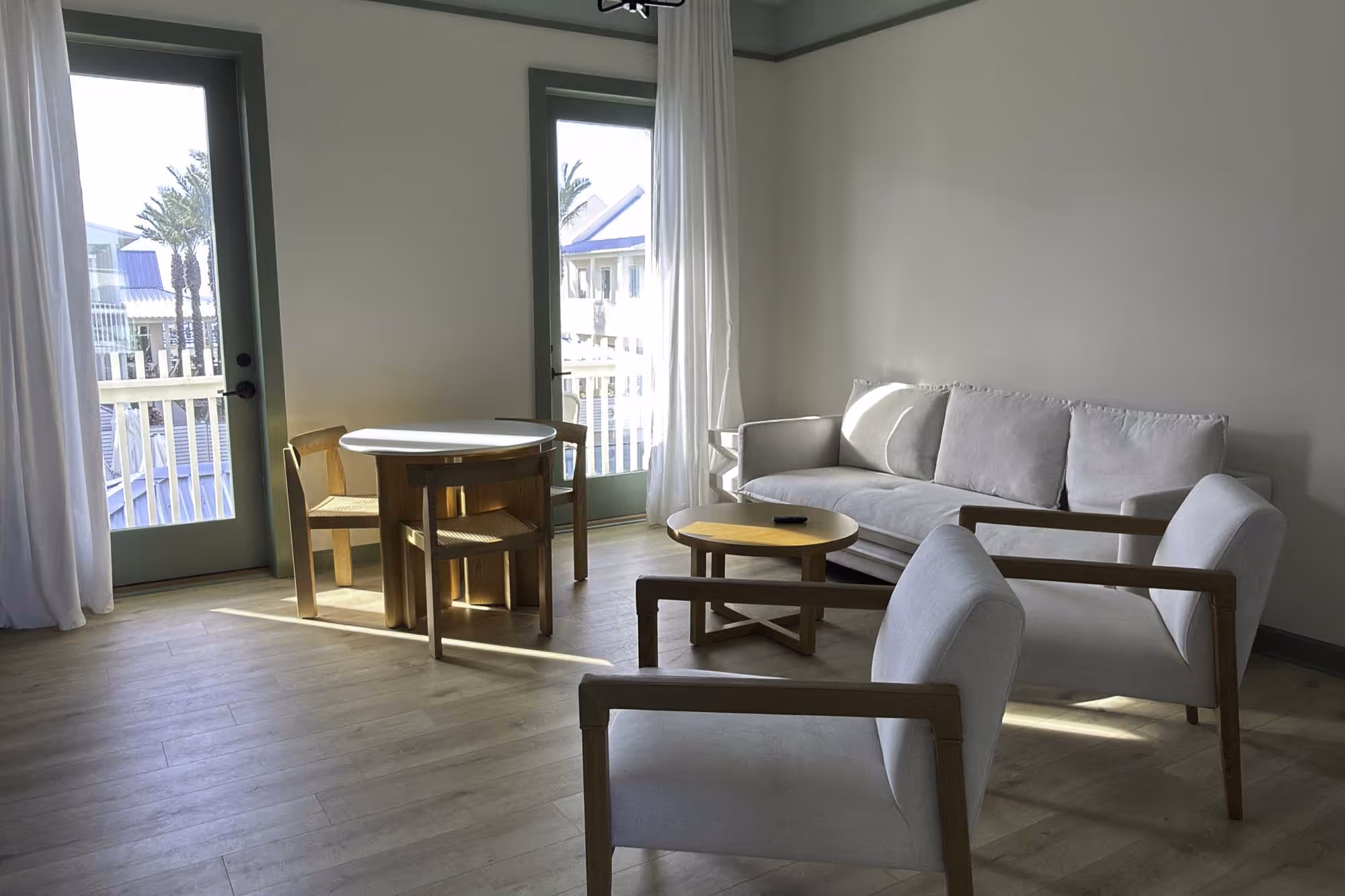 Bright living room with light wood flooring, white sofa, two wooden armchairs with white cushions, round wooden coffee table, and small round dining table with four wooden chairs near two glass doors with white curtains.