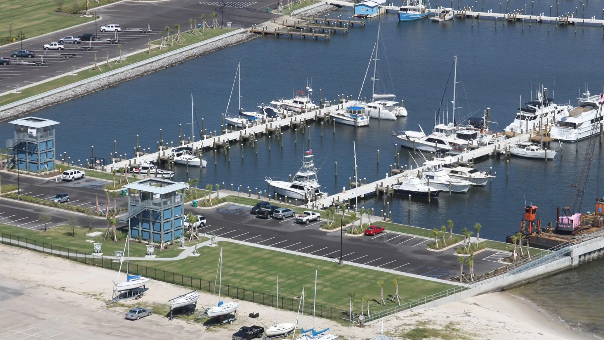 Aerial view of a marina with multiple boats docked alongside piers, two blue observation towers, and parked cars in adjacent lots.