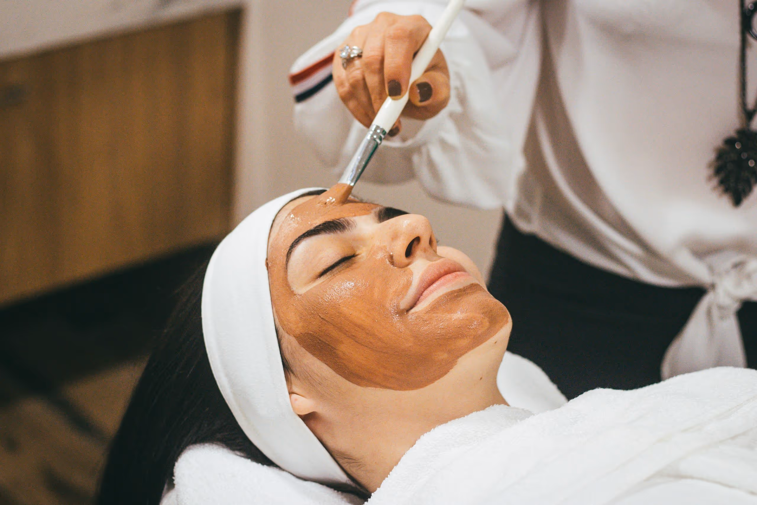 Woman receiving a brown facial mask treatment with a brush while lying down with eyes closed and hair wrapped in a white headband.