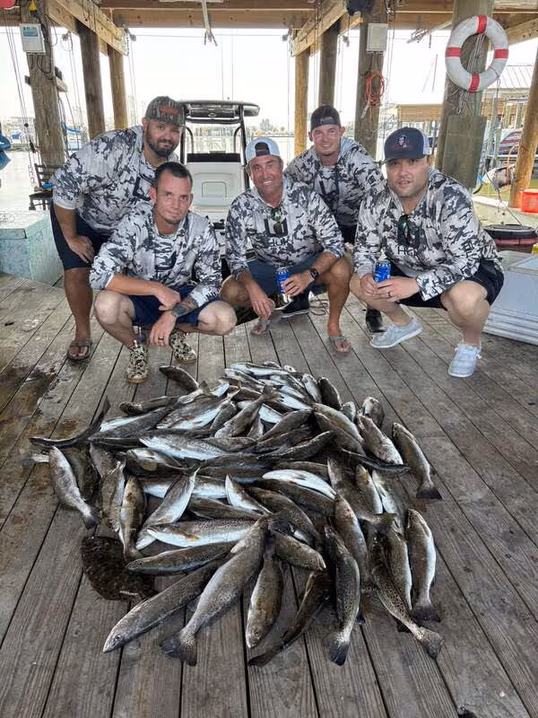 Five men in matching camo shirts crouching behind a large pile of freshly caught fish on a wooden dock under a covered structure.