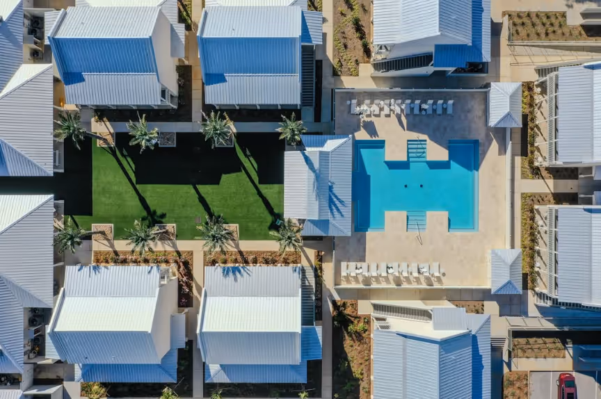 Aerial view of a residential complex with white-roofed buildings surrounding a central swimming pool and a grassy area with palm trees.