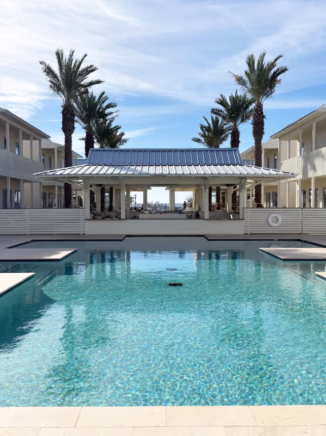 Clear swimming pool in front of modern beachside pavilion flanked by palm trees and two-story buildings.