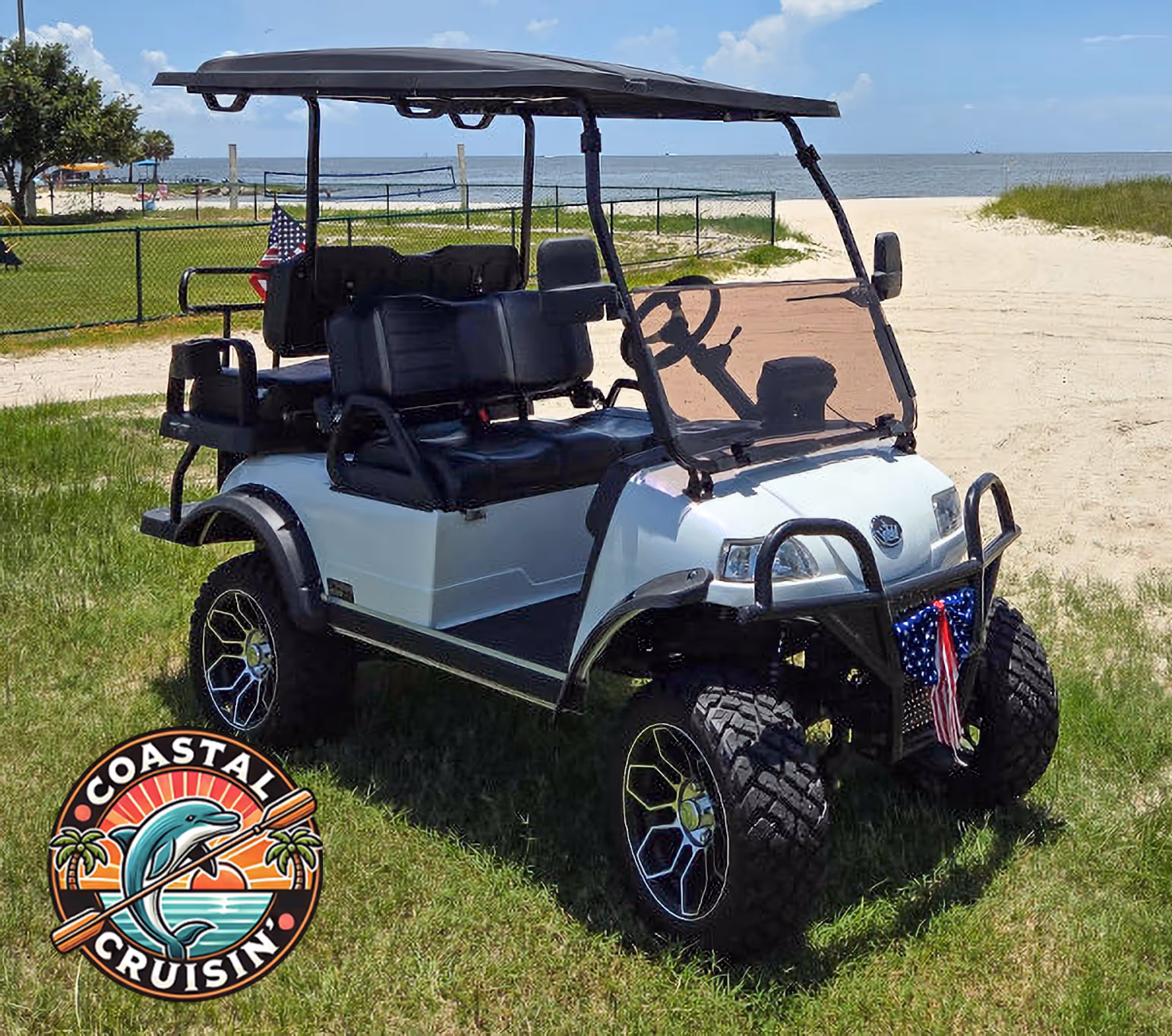 White golf cart with black seats and canopy parked on grass near a sandy beach, decorated with small American flags.