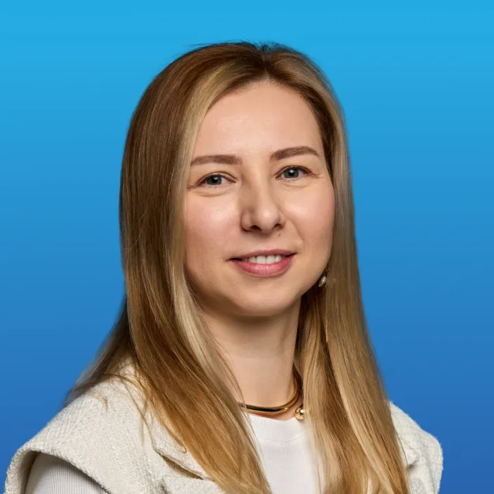 Smiling woman with long blonde hair wearing a white top and textured vest against a blue background.