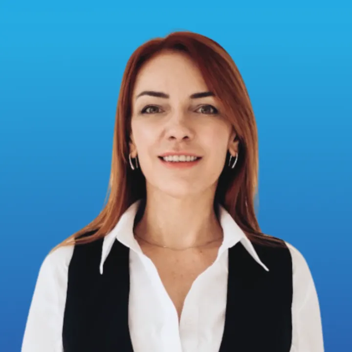 Smiling woman with straight red hair wearing a white shirt and black vest against a blue background.