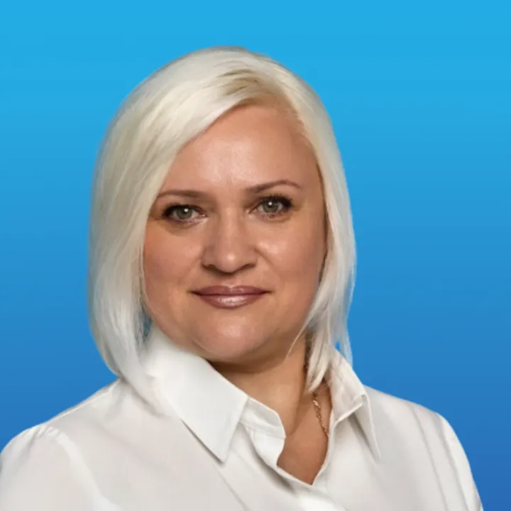 Portrait of a smiling woman with short white hair wearing a white collared shirt against a blue background.