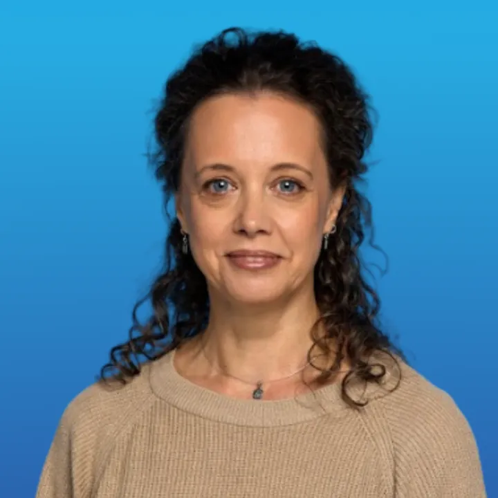 Woman with curly dark hair wearing a beige sweater against a blue background.
