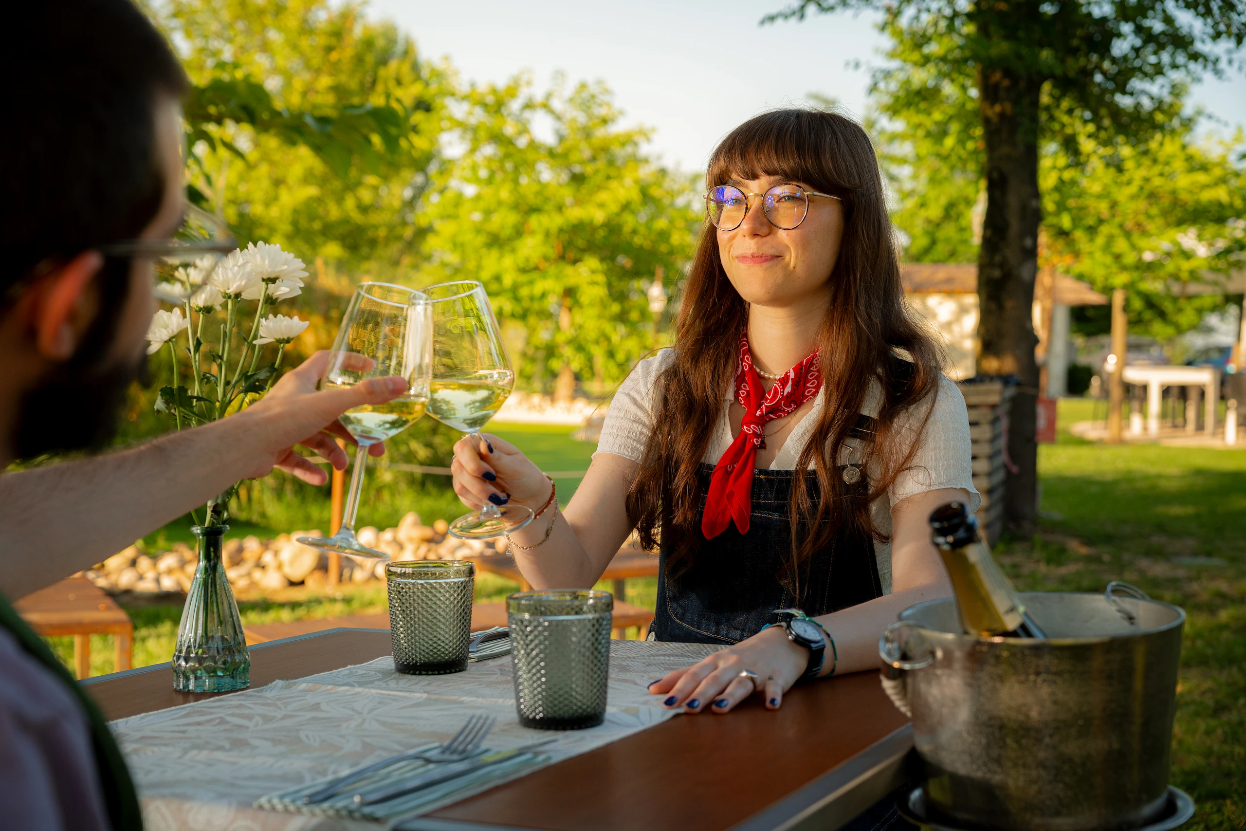Le Bande Ristorante Cavriana, Shooting Studio Looma. Due persone che brindano con calici di vino bianco sedute a un tavolo all'aperto in giardino al tramonto.