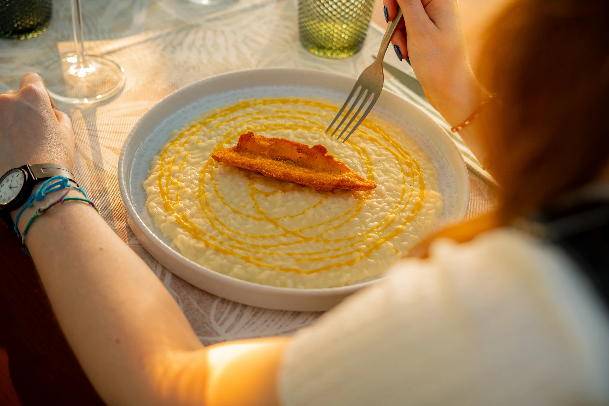 Persona che tiene una forchetta sopra un piatto di risotto cremoso guarnito con un pezzo di pesce fritto e gocce di salsa. Le Bande Ristorante Cavriana, Shooting Studio Looma