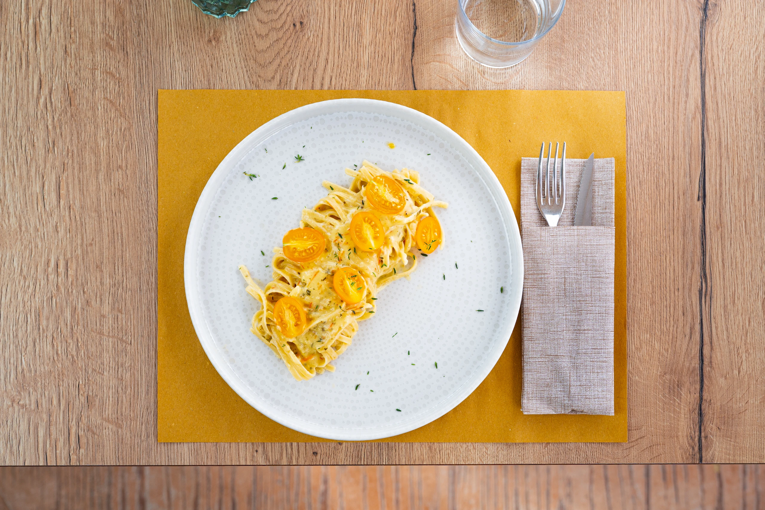 Le Bande Ristorante Cavriana, Shooting Studio Looma, Piatto bianco con pasta condita e pomodorini gialli a metà, posate in un tovagliolo beige su tavolo in legno.