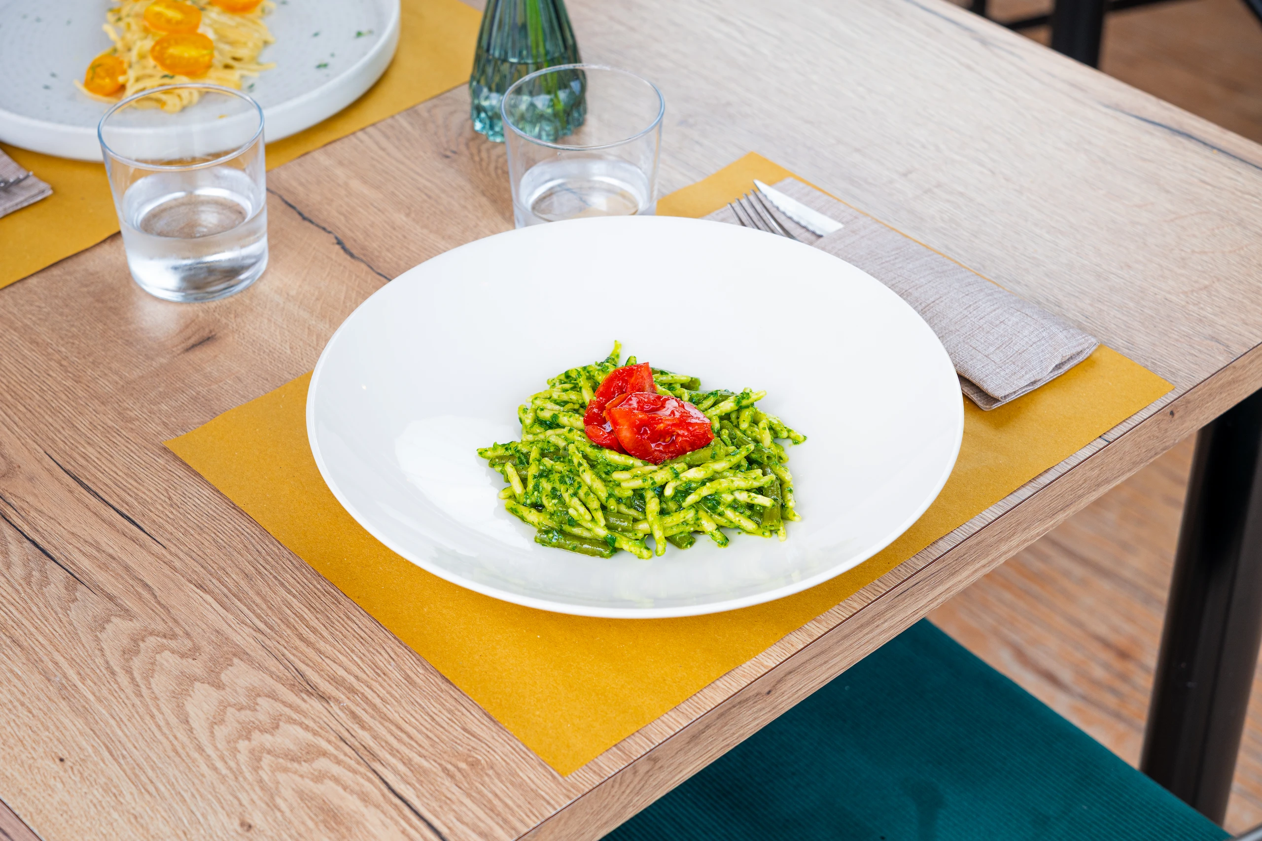 Piatto bianco con pasta con pesto verde e pomodori rossi sopra, servito su tavolo di legno con tovaglietta gialla, bicchiere d'acqua e posate. Le Bande Ristorante Cavriana, Shooting Studio Looma