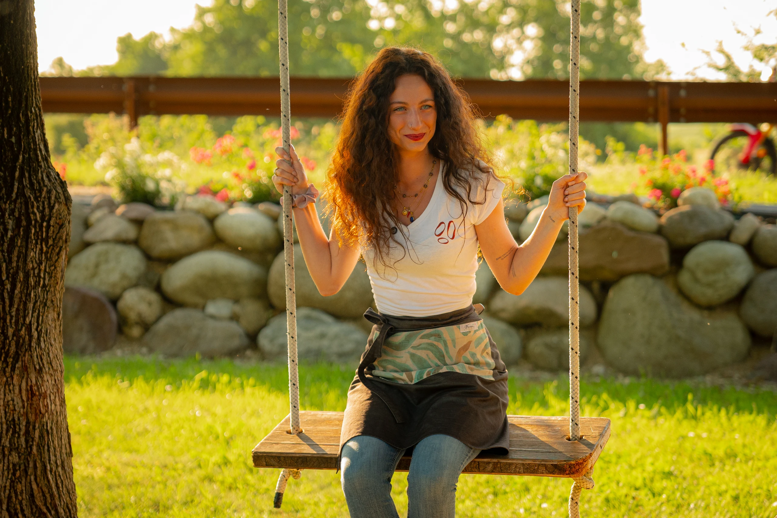 Le Bande Ristorante Cavriana, Shooting Studio Looma. Giovane donna sorridente con capelli ricci castani su un'altalena di legno in un parco soleggiato con pietre e fiori sullo sfondo.