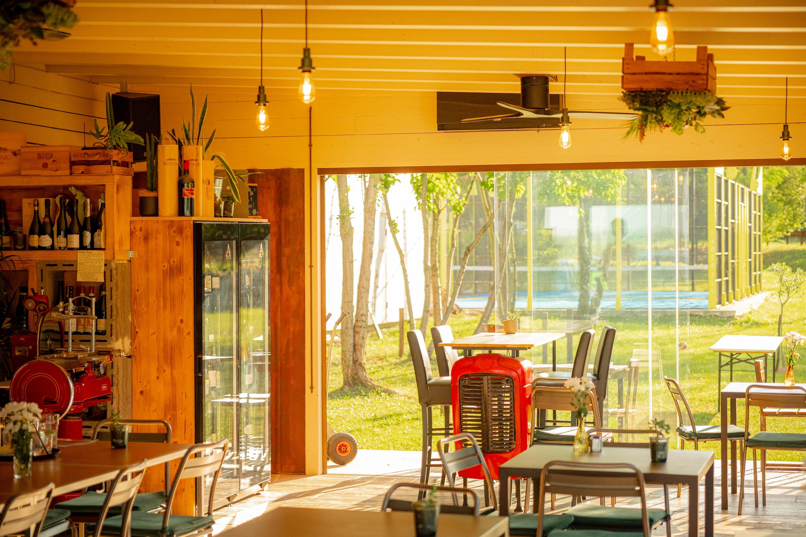 Le Bande Ristorante Cavriana, Shooting Studio Looma.  Interno di un bar luminoso con tavoli, sedie e una vetrina di vini, con vista su un giardino esterno verde.