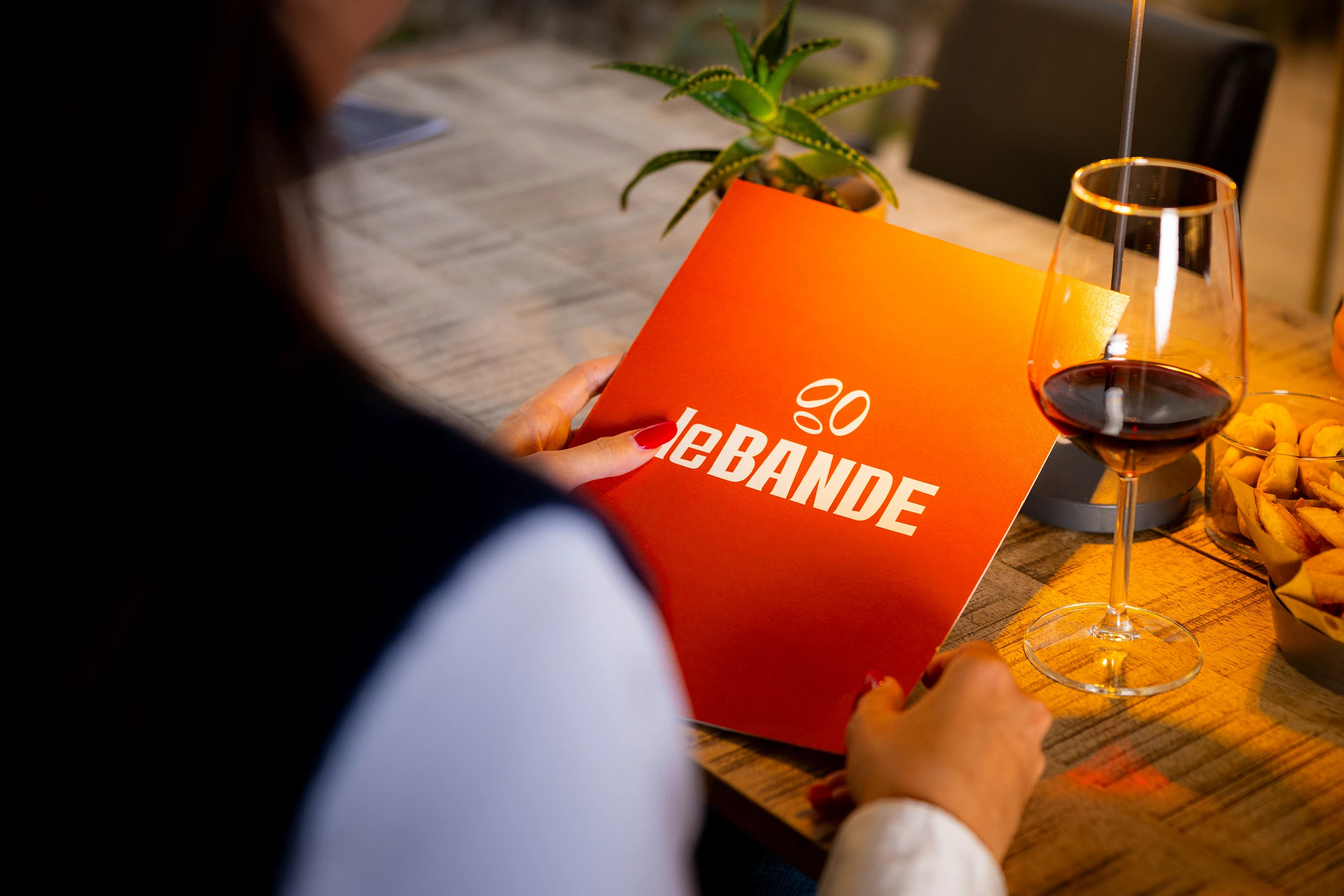Le Bande Ristorante Cavriana, Shooting Studio Looma. Persona che tiene un menu arancione con la scritta LeBANDE, su un tavolo con un bicchiere di vino rosso, una pianta e un contenitore di snack.