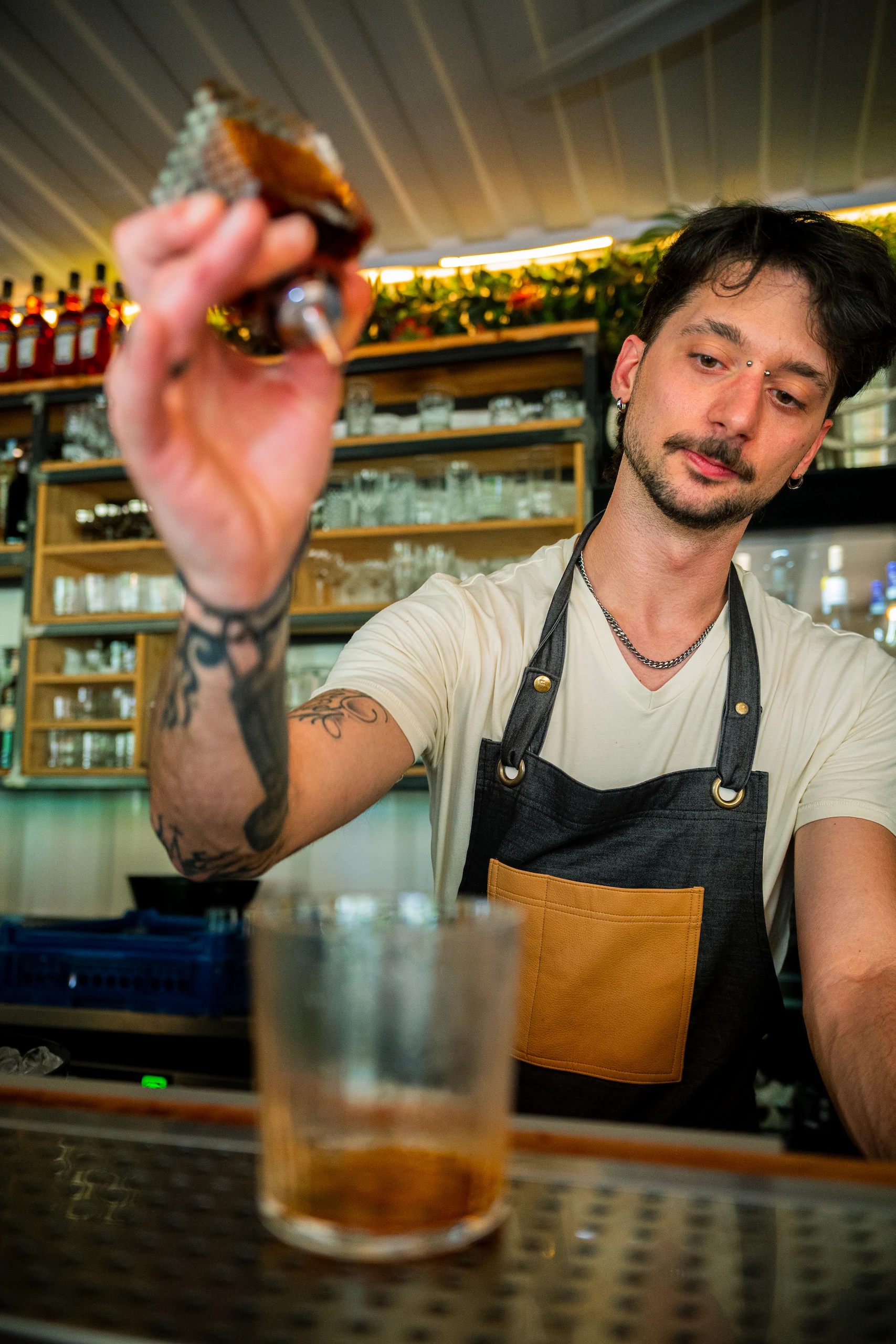 Le Bande Ristorante Cavriana, Shooting Studio Looma. Barman con tatuaggi e grembiule che versa un liquido ambrato in un bicchiere trasparente su un bancone.