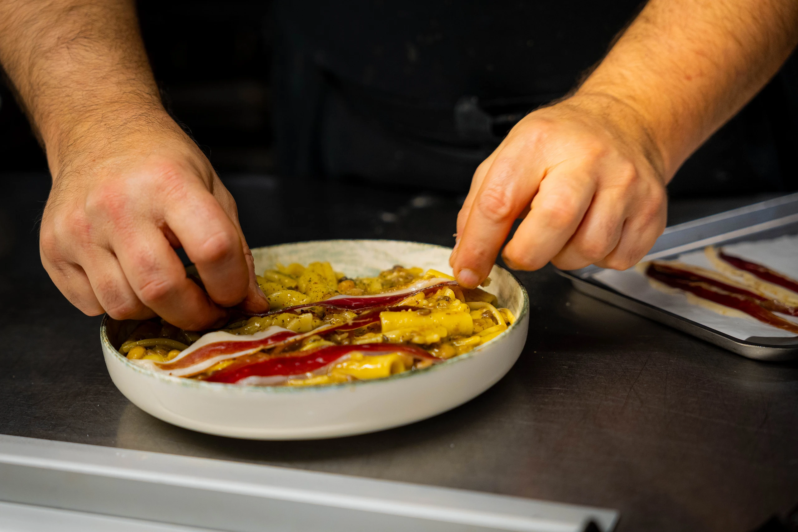 Le Bande Ristorante Cavriana, Shooting Studio Looma. Mani che adagiano fette di pancetta su un piatto di pasta con sugo e semi neri.