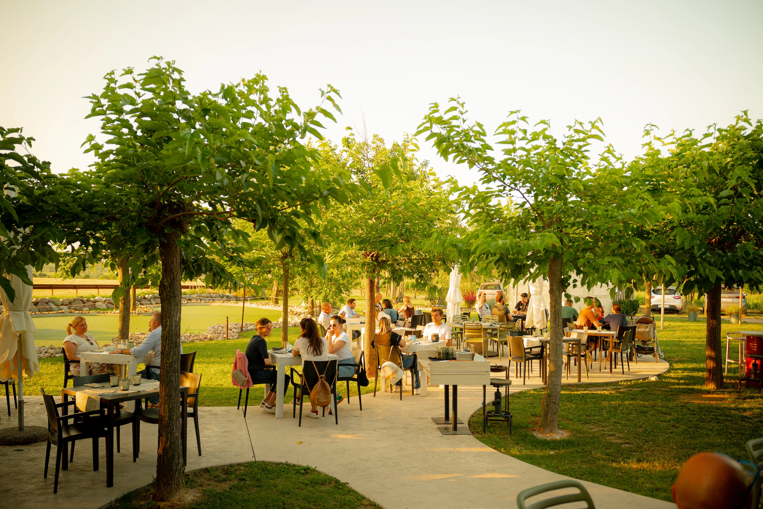 Le Bande Ristorante Cavriana, Shooting Studio Looma. Persone sedute ai tavoli di un ristorante all'aperto in un giardino con alberi verdi e luce calda del tramonto.