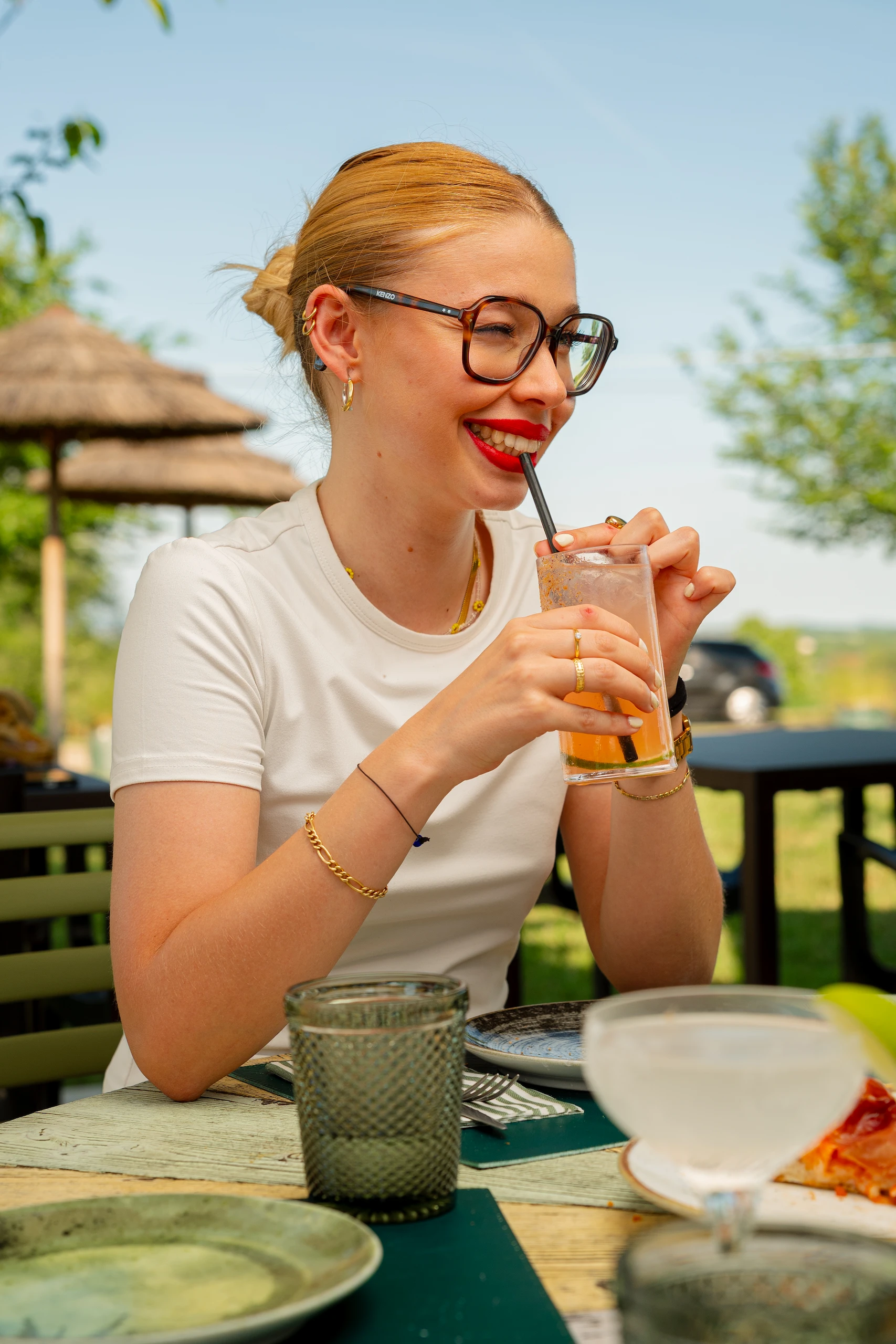 Le Bande Ristorante Cavriana, Shooting Studio Looma. Donna con occhiali grandi e rossetto rosso che sorseggia una bevanda arancione con cannucce in un locale all'aperto.