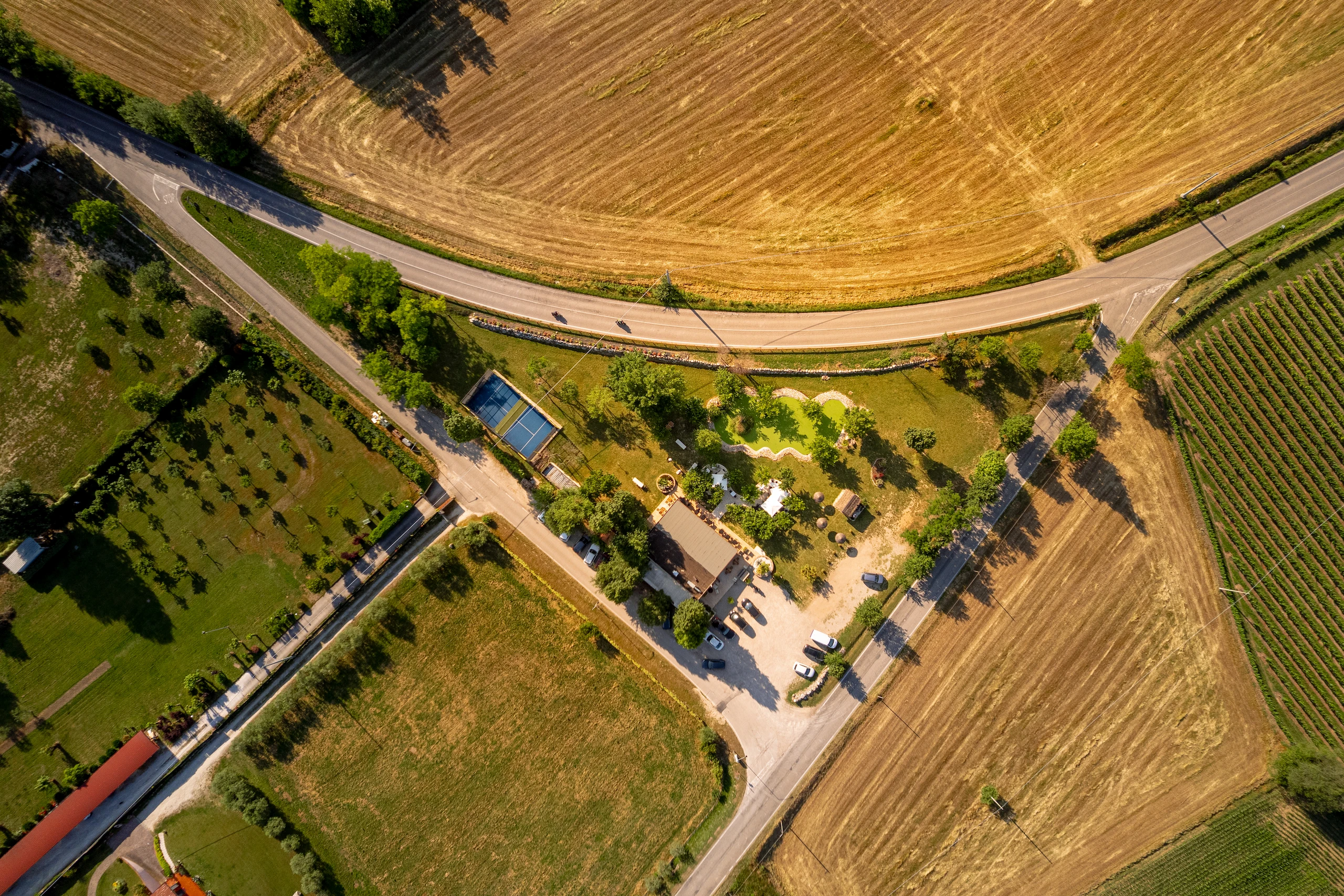 Le Bande Ristorante Cavriana, Shooting Studio Looma. Veduta aerea di una zona rurale con campi gialli e verdi, strade asfaltate, parcheggio con auto, un edificio e un giardino con alberi e sentieri.