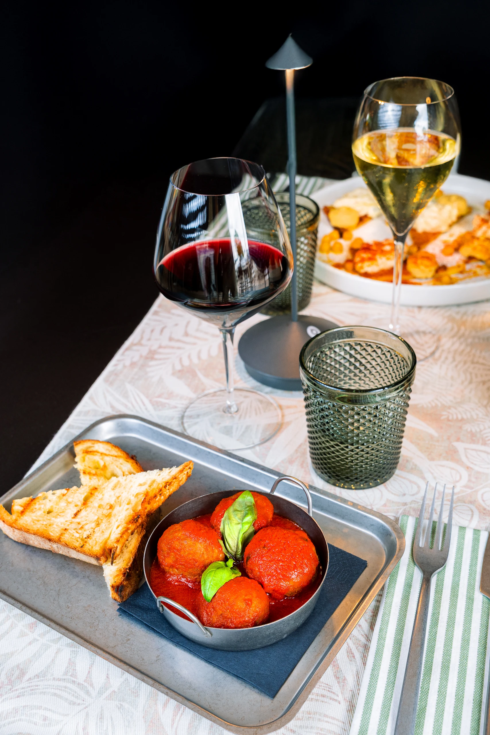 Le Bande Ristorante Cavriana, Shooting Studio Looma. Piatto con polpette di pomodoro guarnite con basilico, accompagnate da fette di pane tostato e due calici di vino rosso e bianco sul tavolo.