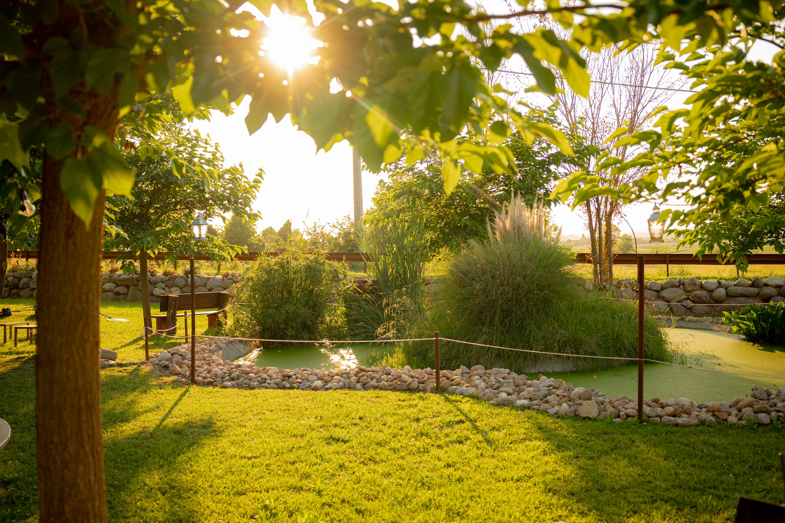 Le Bande Ristorante Cavriana, Shooting Studio Looma. Giardino soleggiato con prato verde, un laghetto circondato da pietre e cespugli, alberi e una panchina di legno.