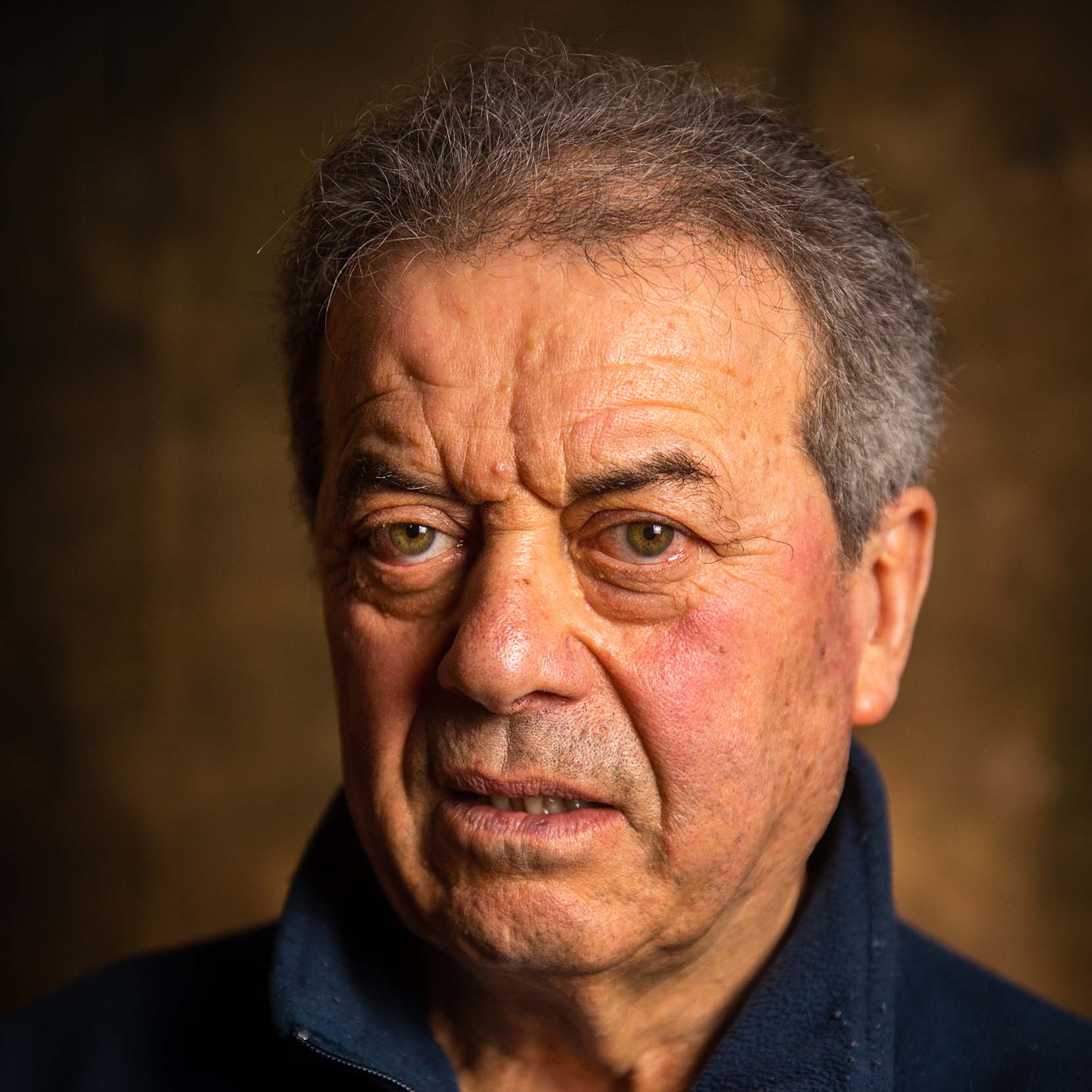 I segni del mestiere, Campagna sociale. Primo piano di un uomo anziano con capelli grigi, espressione seria e occhi verdi su sfondo sfocato marrone.