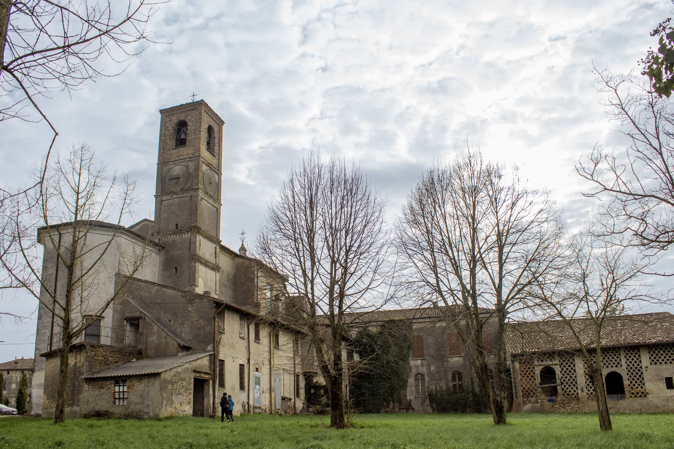 Un paese nuovo, Ceresara, Libro. Edificio storico con torre campanaria in pietra, alberi senza foglie e prato verde, con due persone vicino all'ingresso.