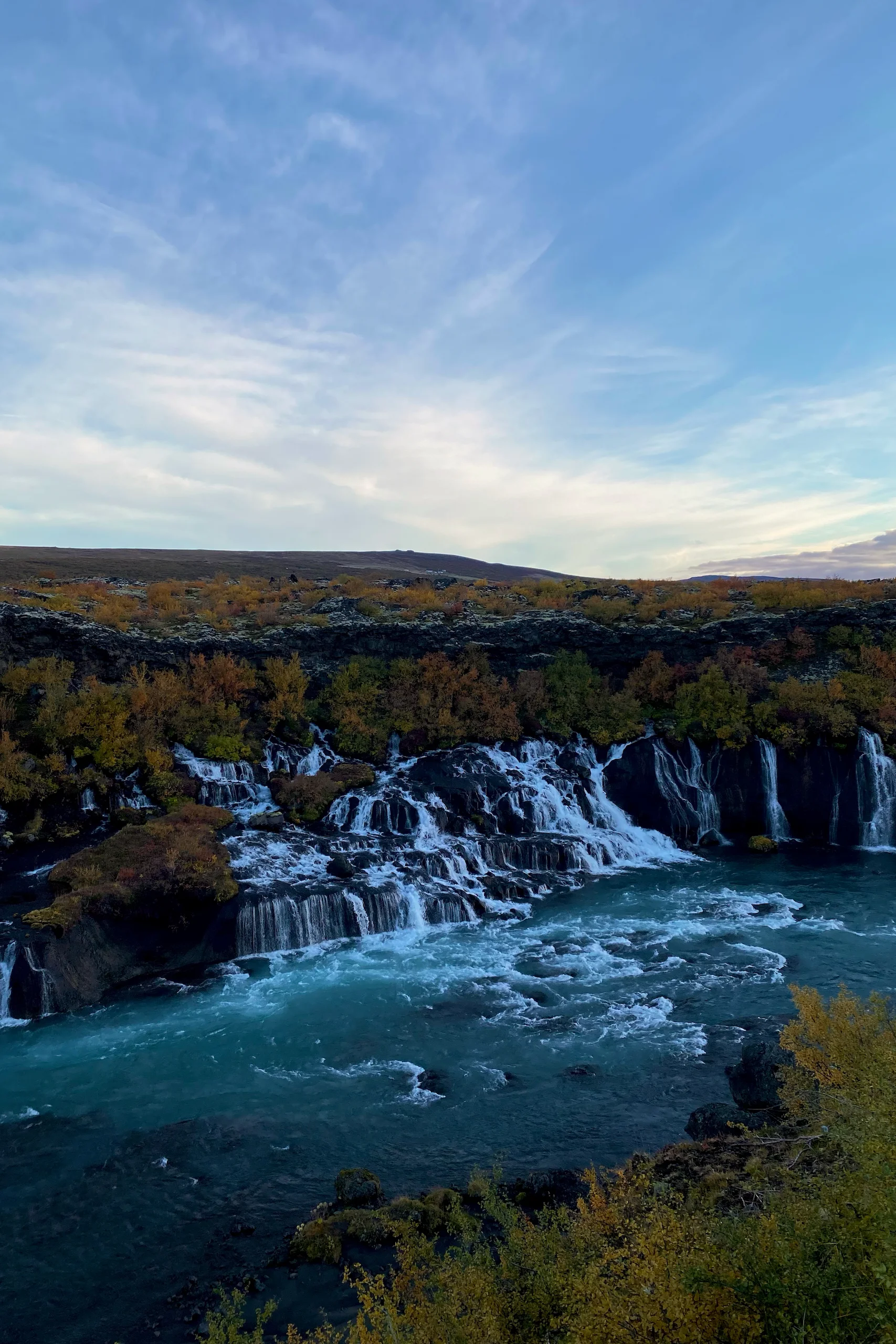 Islanda in auto, libro, Daniel Cortese. Una cascata con numerosi corsi d'acqua che scendono su rocce scure circondate da alberi con foglie autunnali e un fiume turchese in primo piano sotto un cielo azzurro parzialmente nuvoloso.