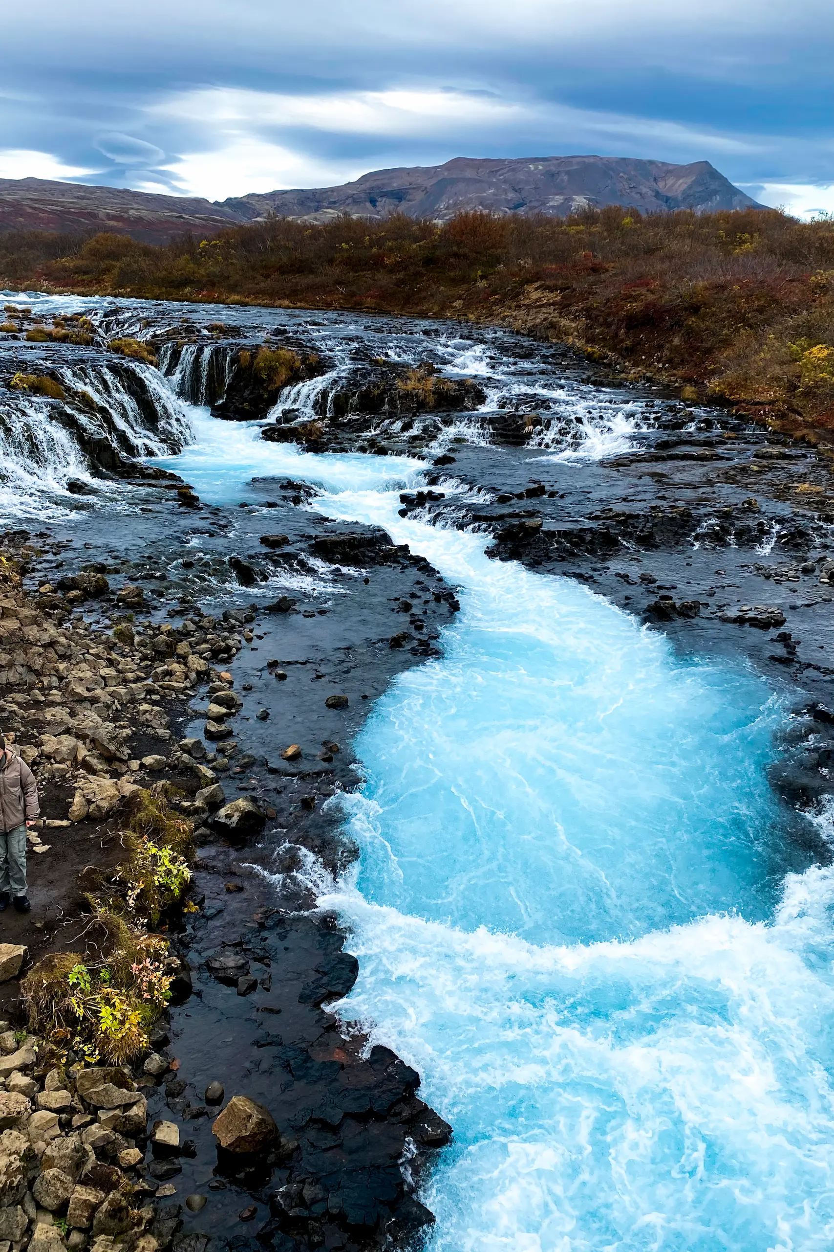 Islanda in auto, libro, Daniel Cortese. Cascata di acqua blu brillante che scorre tra rocce scure con colline e cielo nuvoloso sullo sfondo.