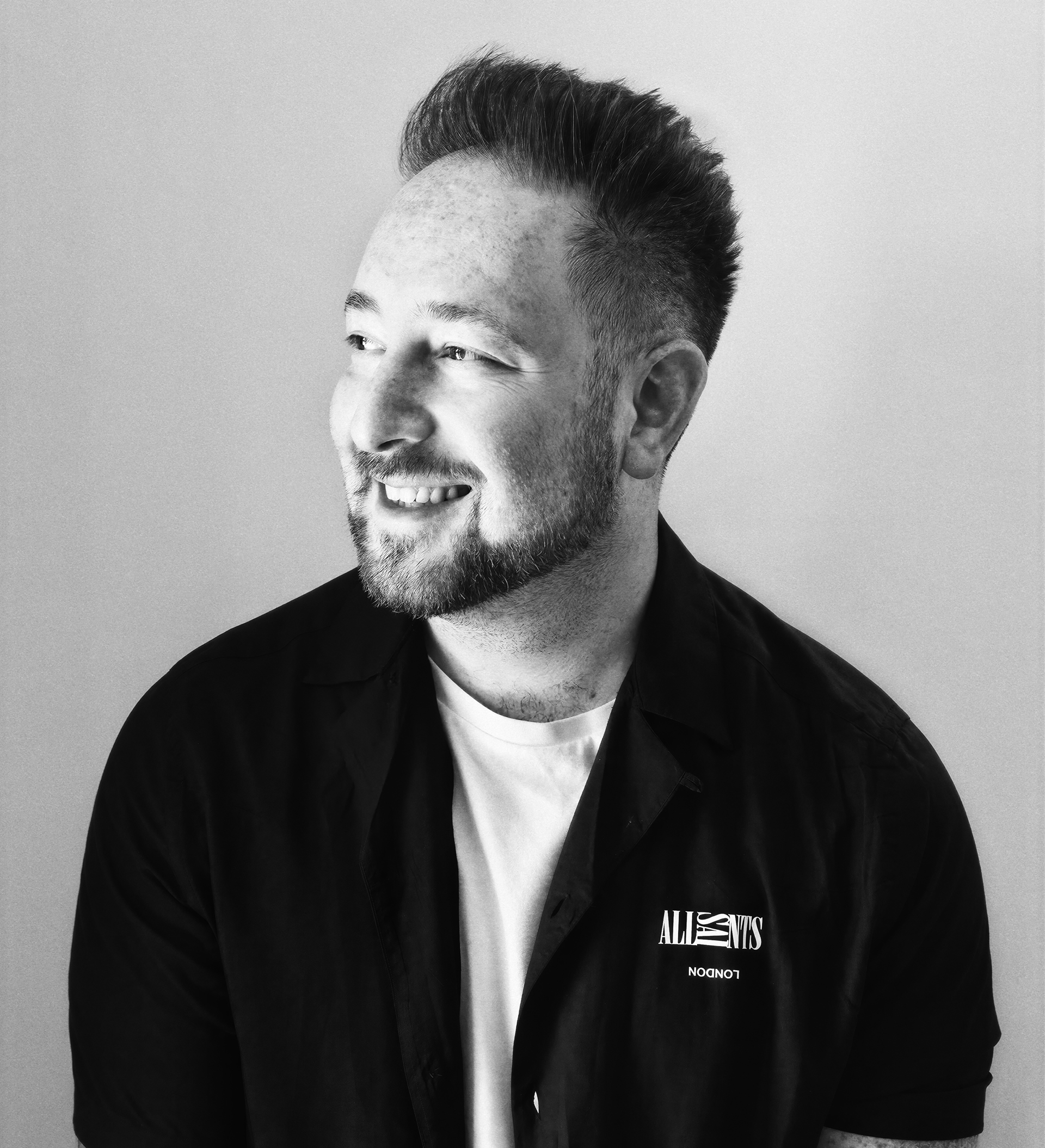 Bearded man with folded arms wearing a denim jacket and black shirt against plain wall.