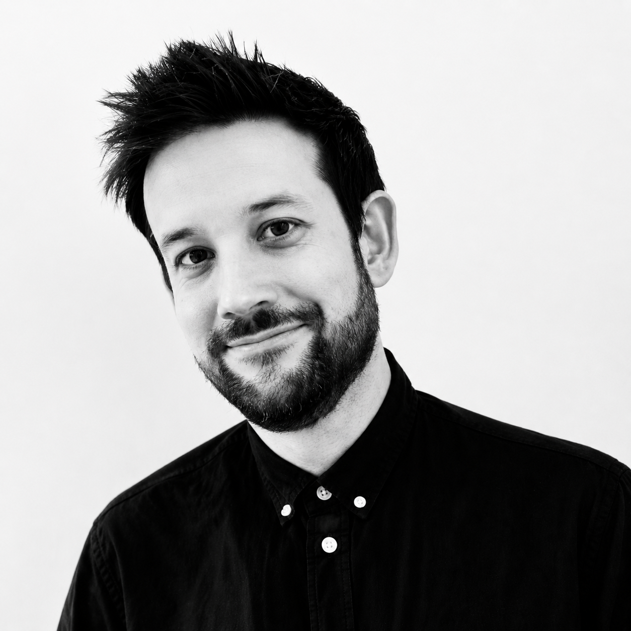 Smiling man with dark hair and beard wearing a black button-up shirt against a white background.