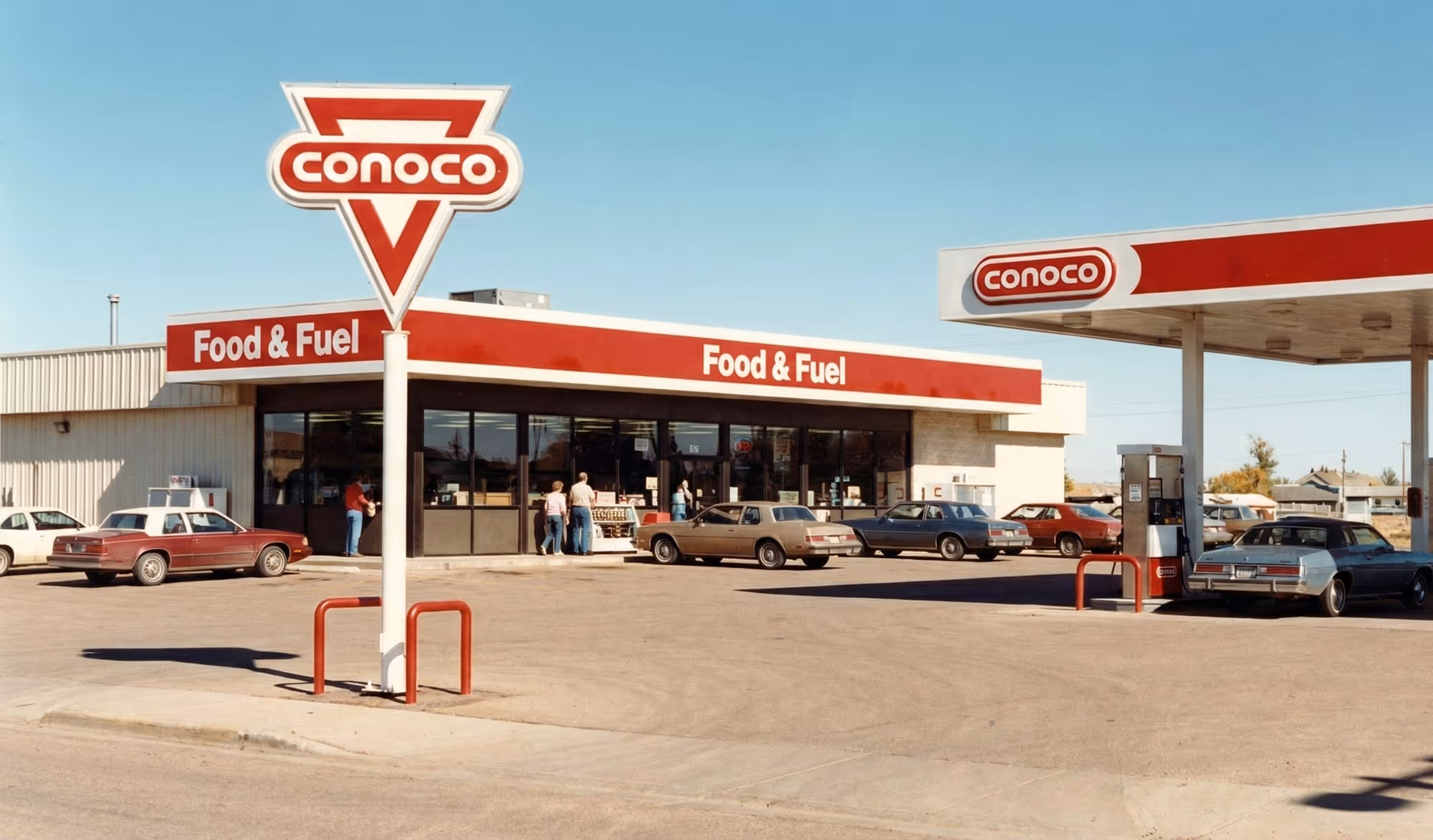 Conoco gas station with food and fuel store, several vintage cars parked outside under clear sky.