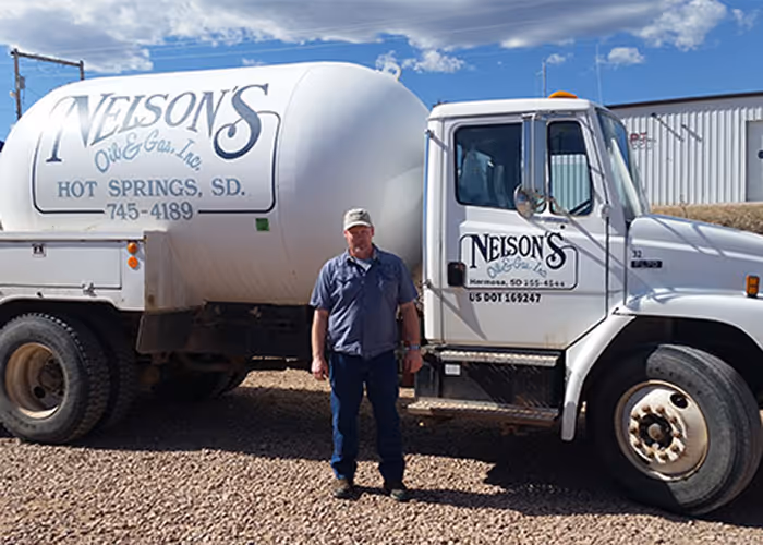Man standing in front of a white Nelson's Oil & Gas propane truck on a gravel surface under a blue sky with clouds.