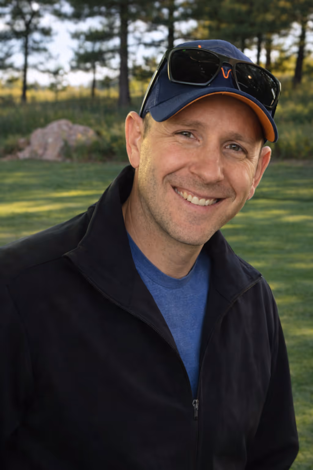 Smiling man wearing a navy cap with sunglasses on top, a black jacket, and a blue shirt standing outdoors with trees and grass in the background.