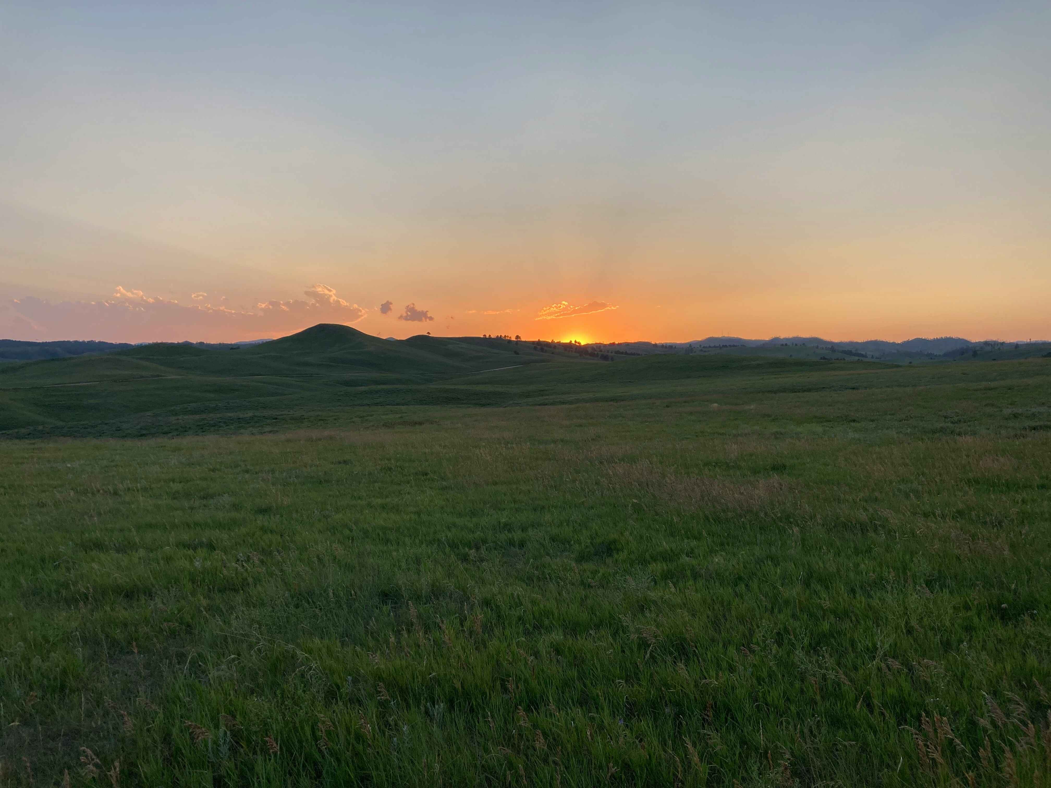Sunset over rolling green hills with a clear sky transitioning from orange to pale blue.