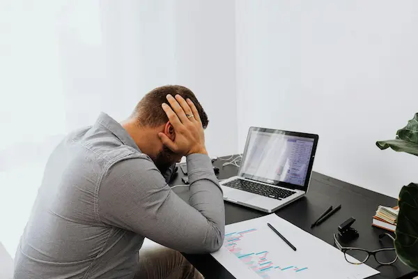 Homme assis à un bureau, tenant sa tête avec les mains, visiblement stressé devant un ordinateur portable et des graphiques papier.