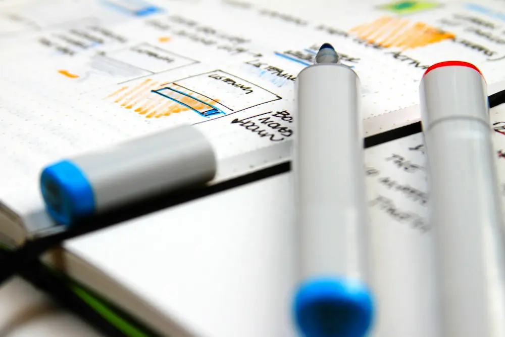 Stylos marqueurs bleus et rouges posés sur un carnet avec des notes manuscrites et des croquis.