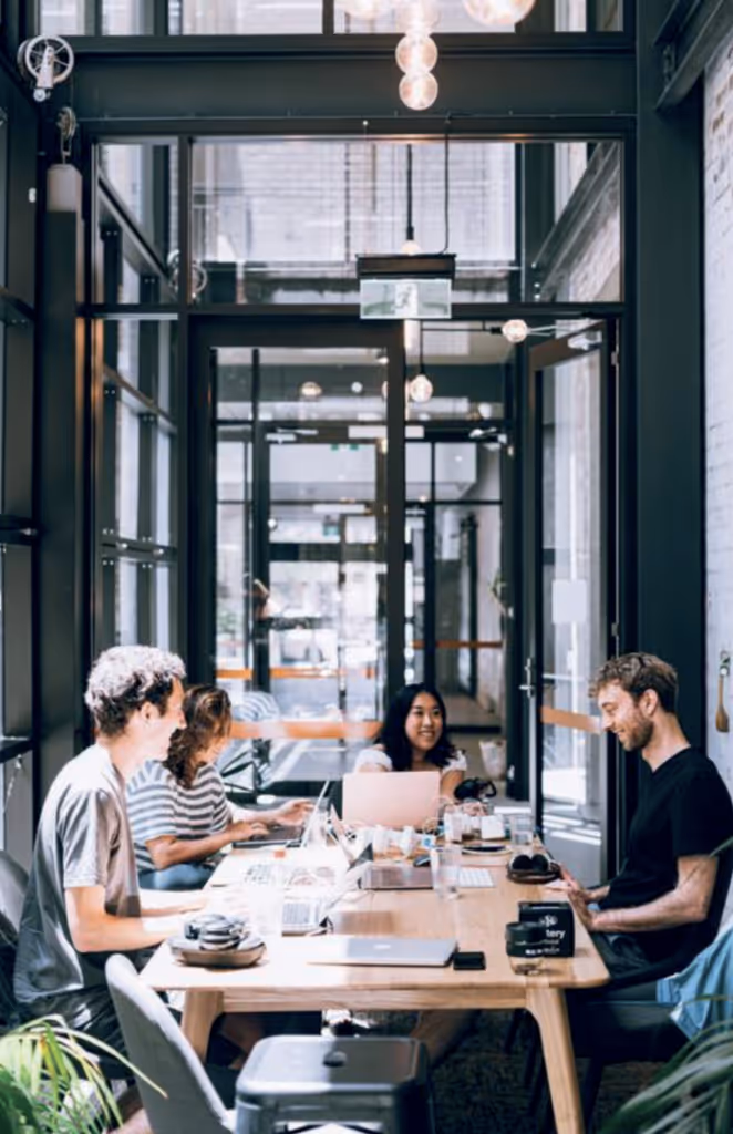 Four people sitting around a wooden table in a modern glass-walled office, working on laptops and smiling.