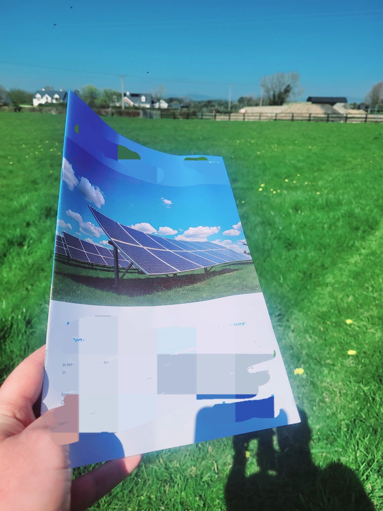 Hand holding a brochure featuring solar panels against a green field and blue sky background.