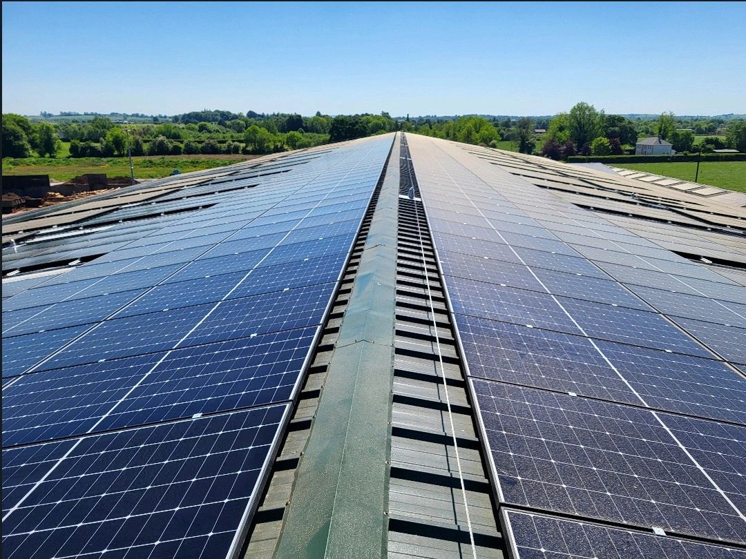 Rows of solar panels installed on a sloped rooftop under a clear blue sky with green fields and trees in the background.