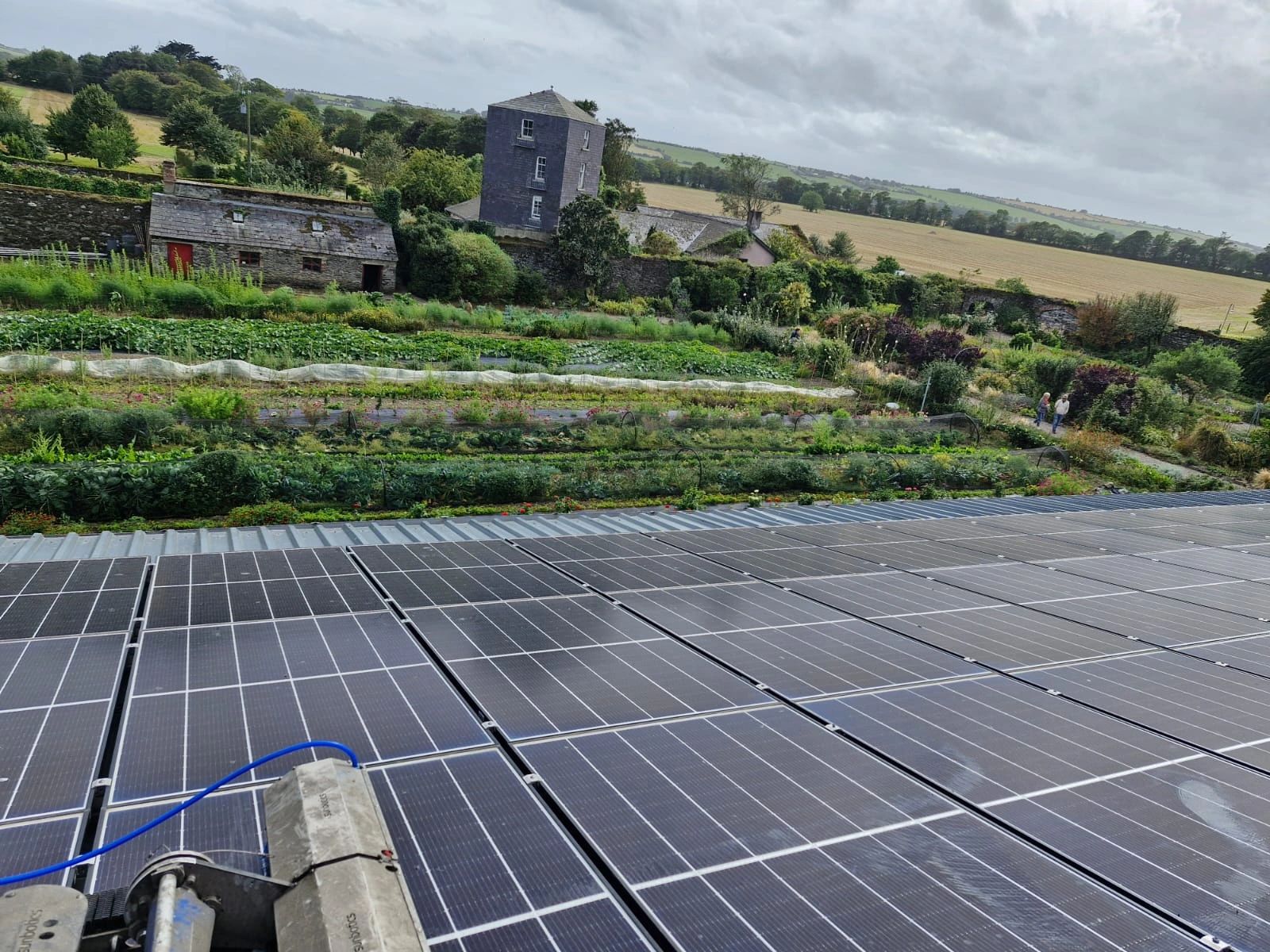Solar panels installed on a rooftop overlooking a garden with rows of plants, old stone buildings, and fields under a cloudy sky.