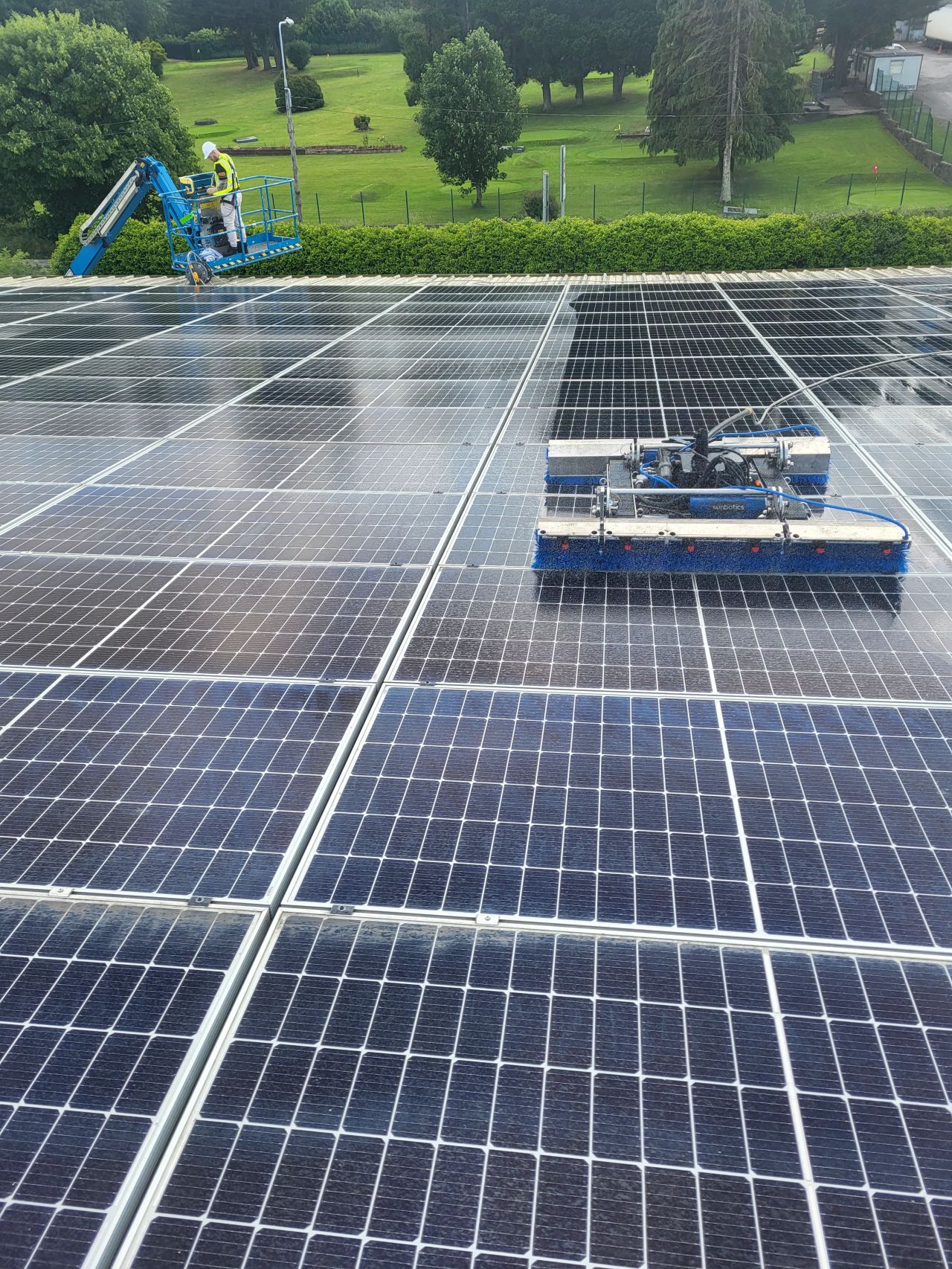 Solar panel array being cleaned by a robotic cleaning machine and a worker on a hydraulic lift in a green outdoor area.