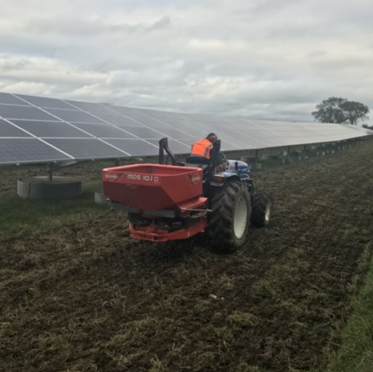 Person driving a tractor with a red fertilizer spreader across a plowed field next to large solar panels under a cloudy sky.